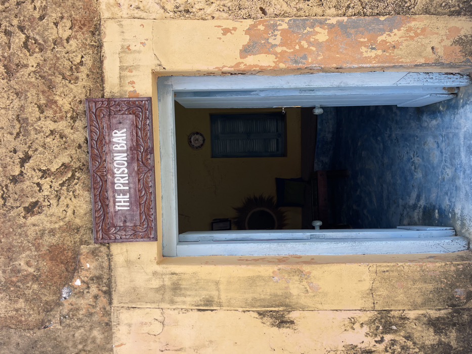An exterior view shows the entrance to "THE PRISON BAR" in Bububu, Tanzania. The building features a heavily weathered, textured yellow facade with significant discoloration, peeling paint, and dark stains, particularly noticeable on the upper section and along the right door frame. Above the doorway, a carved wooden sign with a reddish-brown frame and white lettering clearly reads "THE PRISON BAR." The doorway is framed by light yellow stucco and features a set of light blue, possibly wooden, double doors that are open inward, revealing a dimly lit interior. The doors have simple, clear glass doorknobs. Inside, the walls appear to be painted yellow, and the floor is a dark blue, showing signs of wear. Visible interior elements include a window with closed blue louvered shutters on the back wall and a circular decorative object, possibly a mirror or sunburst art piece, on the left wall. A dark-colored bench or low seating arrangement is partially visible to the left of the window. No individuals or active interactions are depicted in the scene.