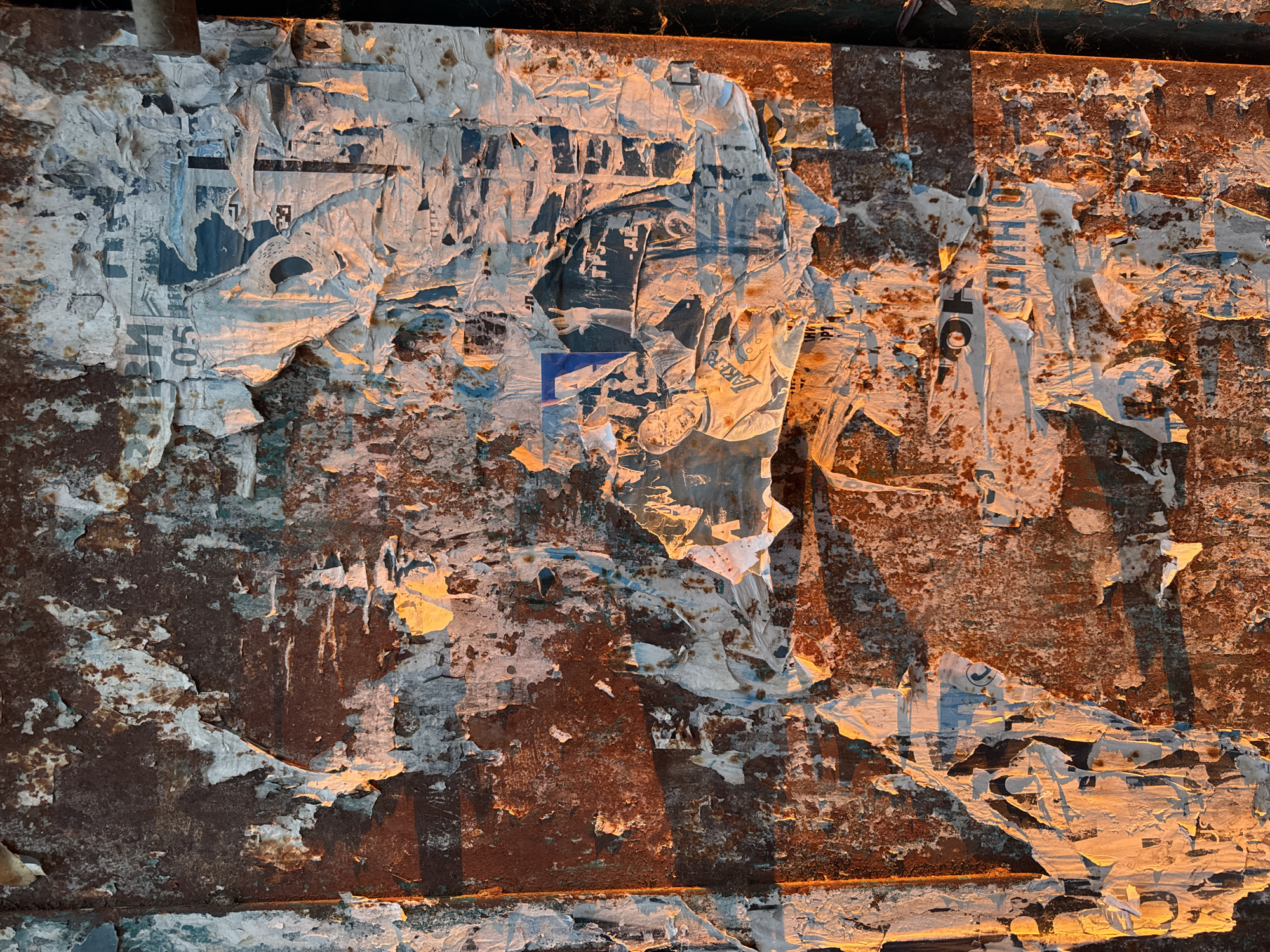 A close-up, vertical shot depicts a heavily textured, dilapidated surface, likely a metal panel or utility box, exhibiting advanced stages of rust and overlaid with multiple layers of torn and peeling paper posters. The predominant underlying material displays deep reddish-brown and orange rust, with remnants of faded light blue-green paint visible beneath. Numerous fragments of white and off-white paper posters are adhered and delaminated across the surface, some crumpled and curling.
Discernible textual elements on the posters include fragmented Cyrillic script, such as the letters "ОН" and "ДИН" in the lower central portion, and the number "05" in the upper central area. A distinct graphic in the upper-right quadrant features a fragment of a basketball player's jersey with the word "LAKERS" printed in yellow, and a partial image of a human hand gesturing upward is visible nearby.
The scene is illuminated by strong, warm, low-angle light originating from the left, casting golden-orange hues on the raised textures and leftward surfaces, while creating pronounced shadows that accentuate the depth and unevenness of the materials. No individuals are present. The environmental context, characterized by weathered urban materials and Cyrillic script, is consistent with a public outdoor setting within the city of Sofia, Bulgaria.