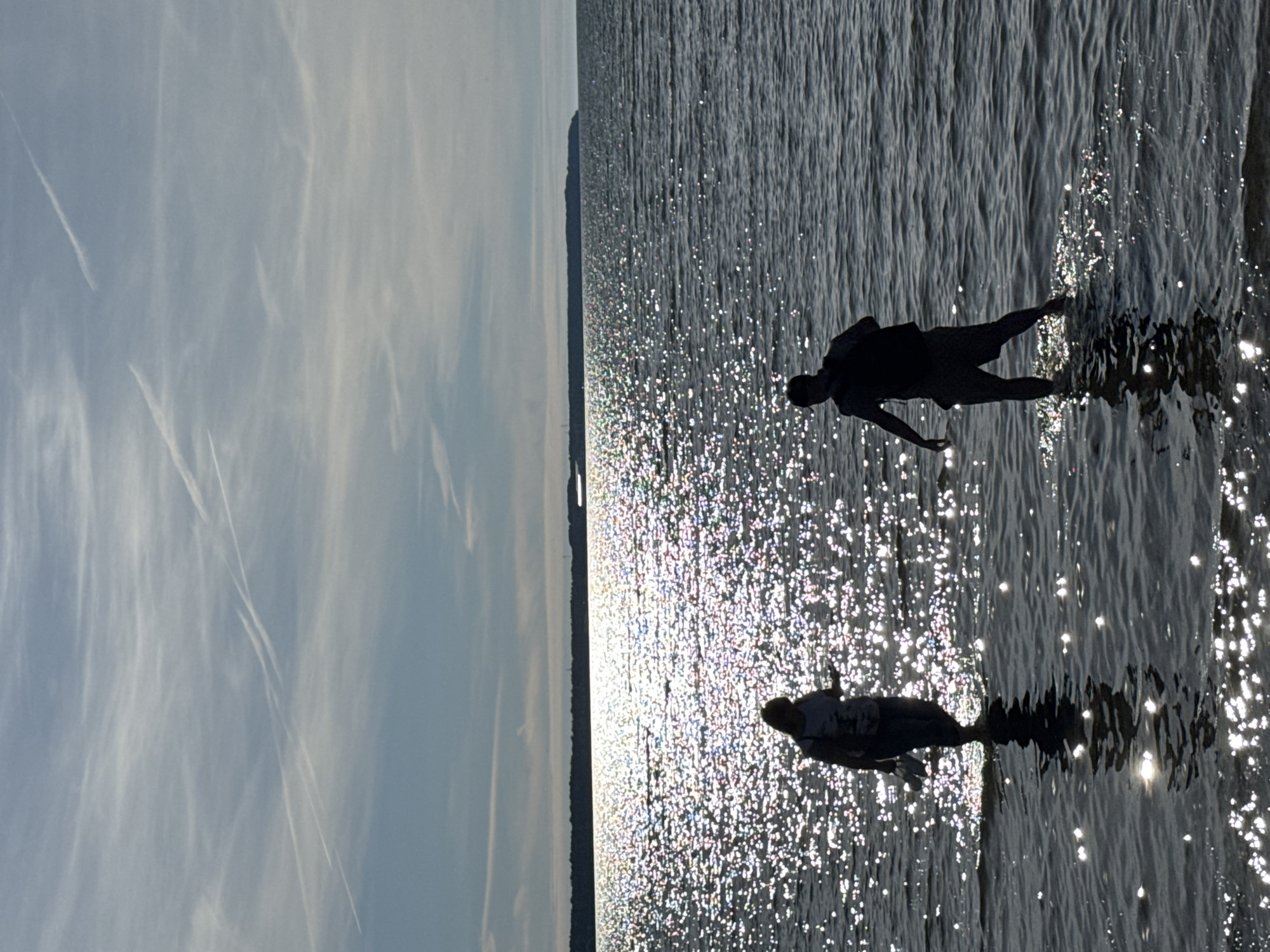 The image depicts a serene body of water, likely a bay or sea, with a distant shoreline visible. The sun is high in the sky, casting a brilliant shimmer across the water's surface, creating a dazzling effect. The sky is a pale blue, streaked with wispy white clouds and contrails from aircraft.
In the foreground, two figures are silhouetted against the bright water. On the left, a person is walking into the water, holding what appears to be a pair of shoes in their hand. They are wearing a light-colored top and darker pants. On the right, another person, also silhouetted, stands in the water, facing away from the camera. They are wearing a backpack. Both individuals are walking through shallow water, their reflections visible below them. The overall impression is one of a peaceful outdoor scene, possibly a beach or a waterfront area in Tallinn, Estonia. The specific location within Tallinn is not identifiable from the image alone.