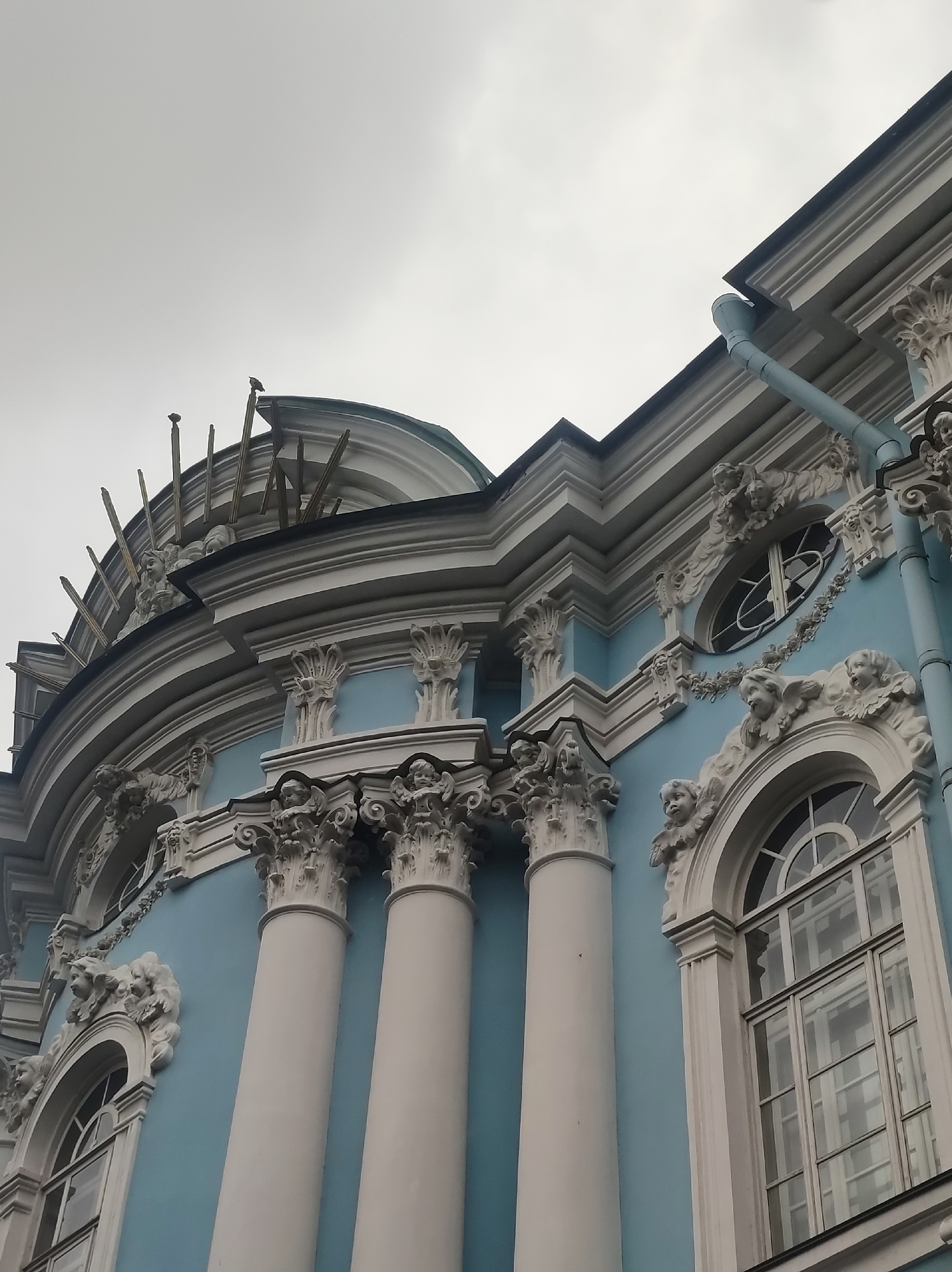 The image shows an upward-angle view of the exterior of the Smolny Cathedral in Saint Petersburg, Russia. The cathedral's distinctive blue walls are adorned with white Baroque-style stucco decorations, including columns, window frames, and cherubic figures. Sunlight reflects off the windows, and a drainpipe is visible along the edge of the roof. The sky is overcast.
