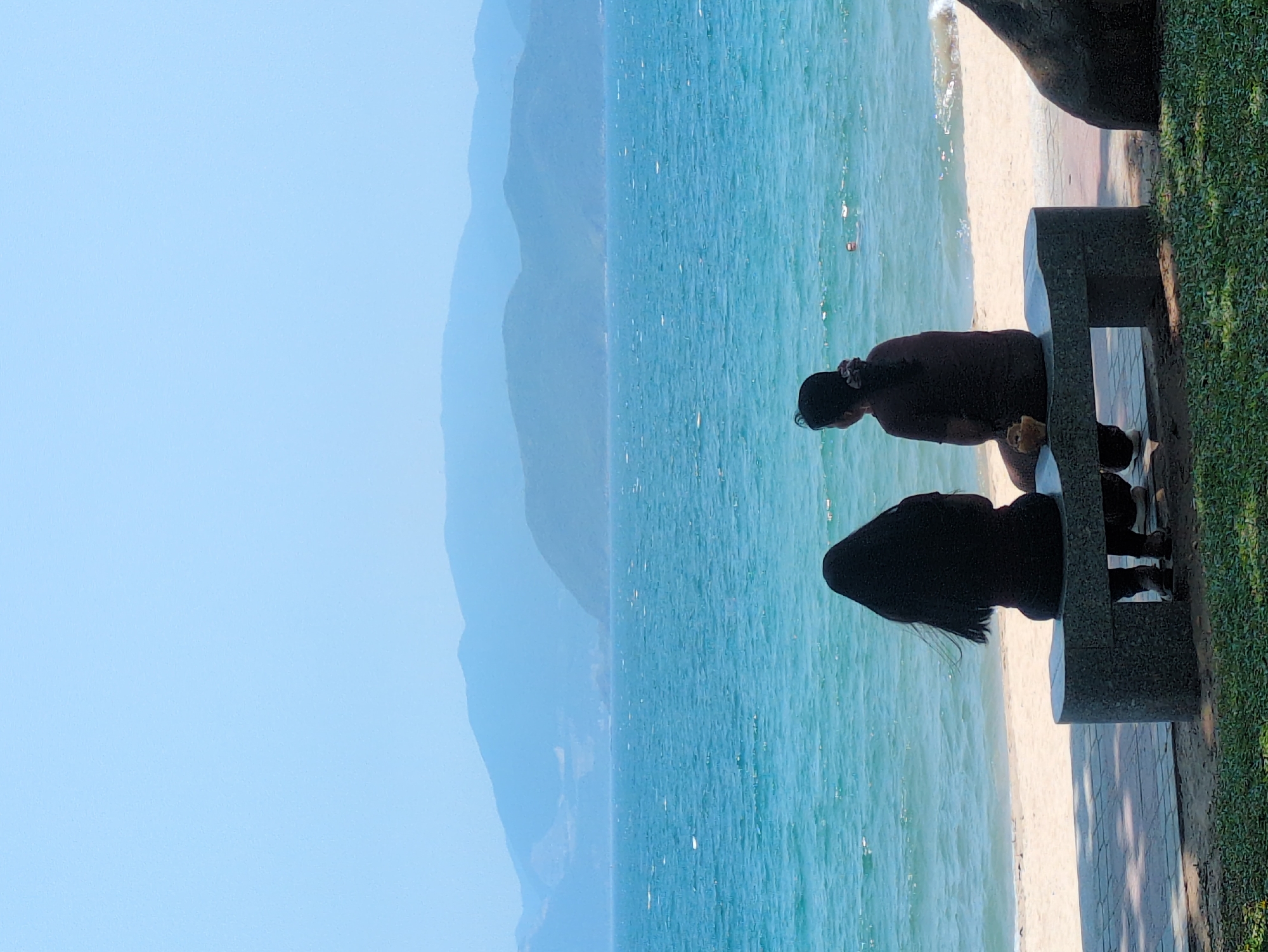 The image depicts two people sitting on a concrete bench facing the beach in Nha Trang, Vietnam. The turquoise ocean stretches out in front of them, with waves gently lapping the shore. In the distance, hazy mountains are visible. The sky is a clear, pale blue. One person is wearing a dark shirt and facing down. The other person has long black hair. The bench is located along a walkway next to a grassy area.