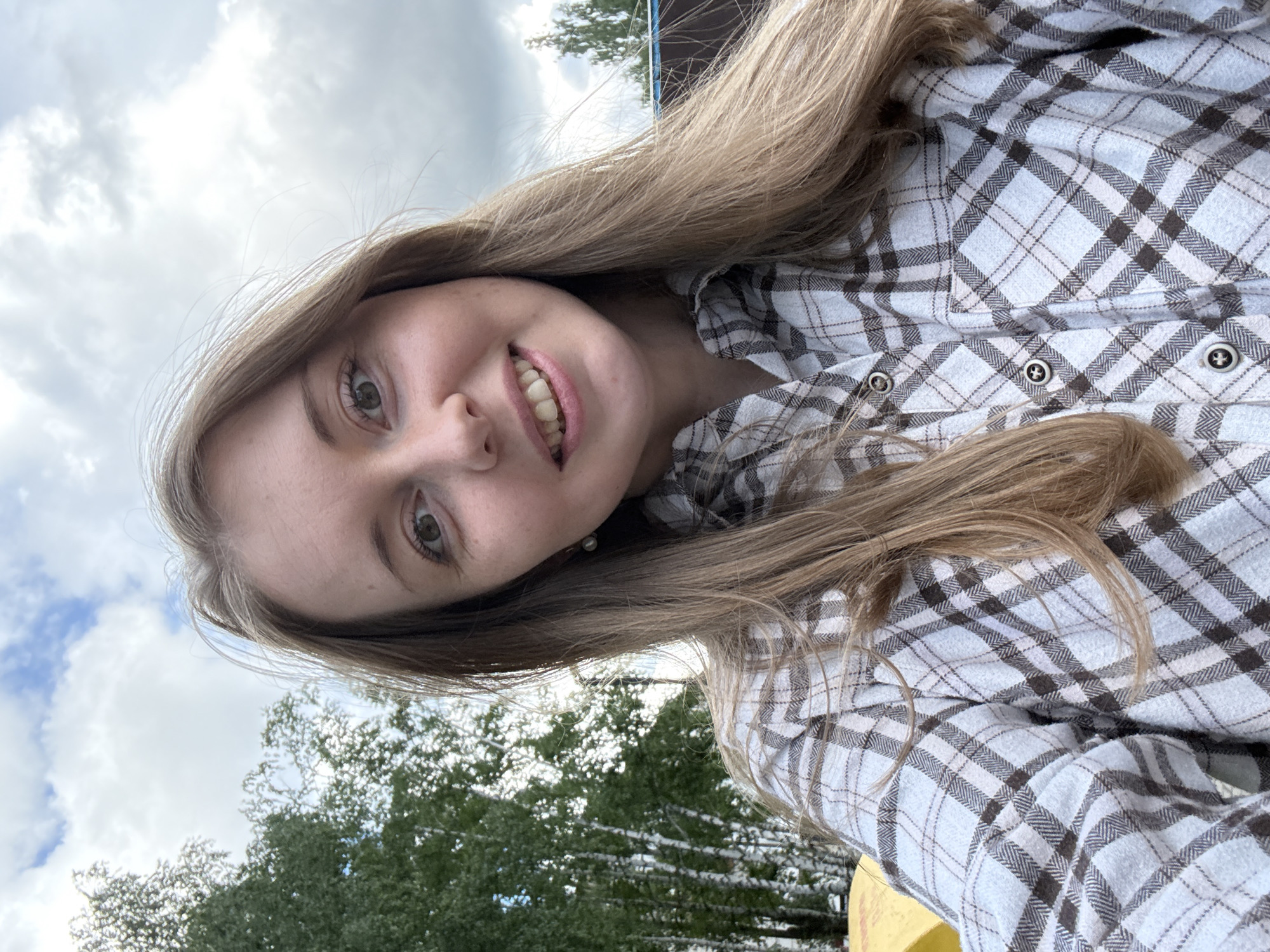 The media file is a rotated selfie of a young woman, captured outdoors during the daytime under a cloudy sky. The woman is smiling broadly, revealing her teeth, and looks directly at the camera with green eyes. Her long, light brown hair falls around her shoulders and across her plaid shirt.
She is wearing a flannel shirt with a pattern of white, light gray, and dark brown checks. Several dark buttons with a lighter cross or star design are visible on the shirt. A small, light-colored stud earring, possibly a pearl, is visible in her right ear. The background shows a significant portion of cloudy sky at the top, transitioning to green trees and foliage below. A hint of a blue structure is also visible in the far background on the right side of the original image. The overall impression is one of contentment or happiness in a natural, open-air setting. No visible text is present in the image.