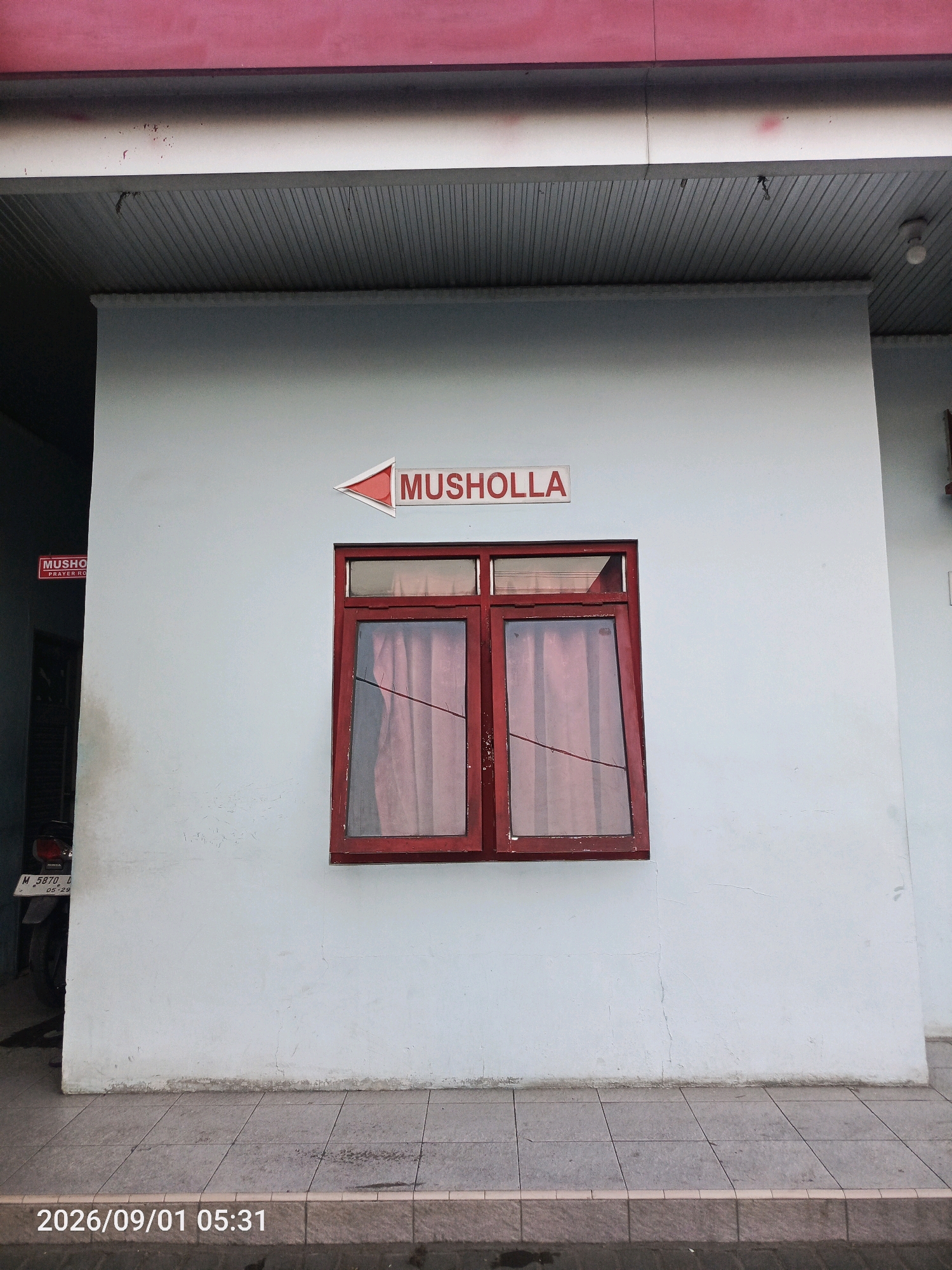 A sign indicating "MUSHOLLA" with an arrow pointing to the right is displayed on a light blue wall. Below the sign is a red-framed window with red curtains. To the left of the window, partially obscured by the building's corner, is a motorcycle with a license plate. The ground in front of the building is tiled. A date and time stamp, "2026/09/01 05:31", is visible at the bottom of the image. This scene is located in Gladak Anyar, Indonesia.
