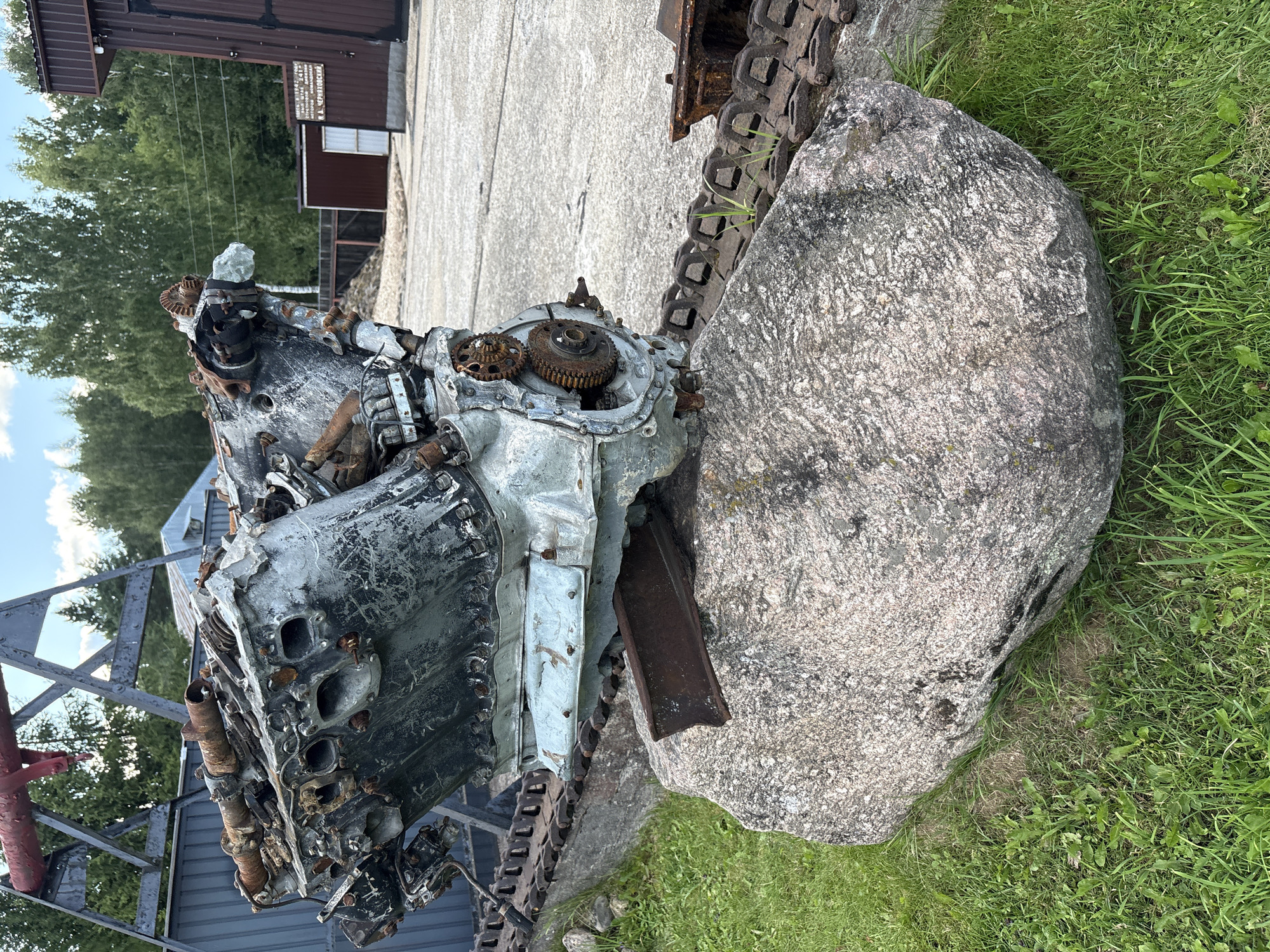 The image shows a large, weathered engine block resting on a rock and a section of tank treads. The engine appears to be a V-engine, possibly from an aircraft or vehicle, with visible rust and damage. It is mounted on a concrete base and partially supported by a metal beam. In the background, there are trees, a corrugated metal building, and a larger industrial-looking structure. The scene is outdoors, with grass growing around the rock and treads. The location is identified as Zhalybnya, Russia, suggesting this may be an exhibit or monument related to industrial or military history. There are no people visible in the image.