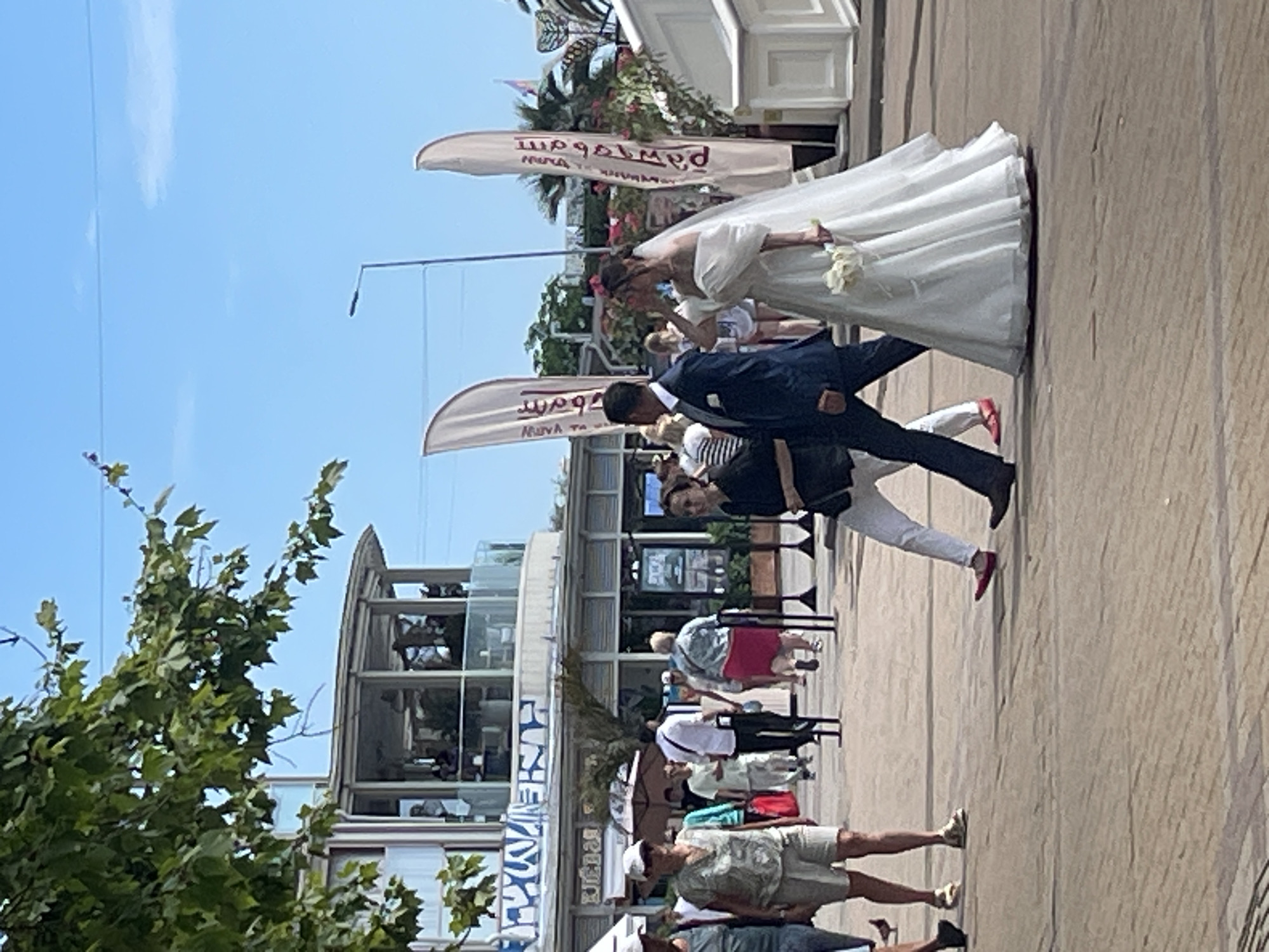 In Sochi, Russia, a bride in a white wedding dress and a groom in a dark suit walk on a paved area. The bride carries a bouquet of white flowers. The groom walks beside her, his left arm extended slightly. Behind them, other pedestrians are visible, some walking in the same direction. In the background, there is a building with a circular glass-enclosed upper level, and two tall, white flags with dark lettering are planted. Trees with green leaves are present on the left side of the frame. The sky is clear and blue.