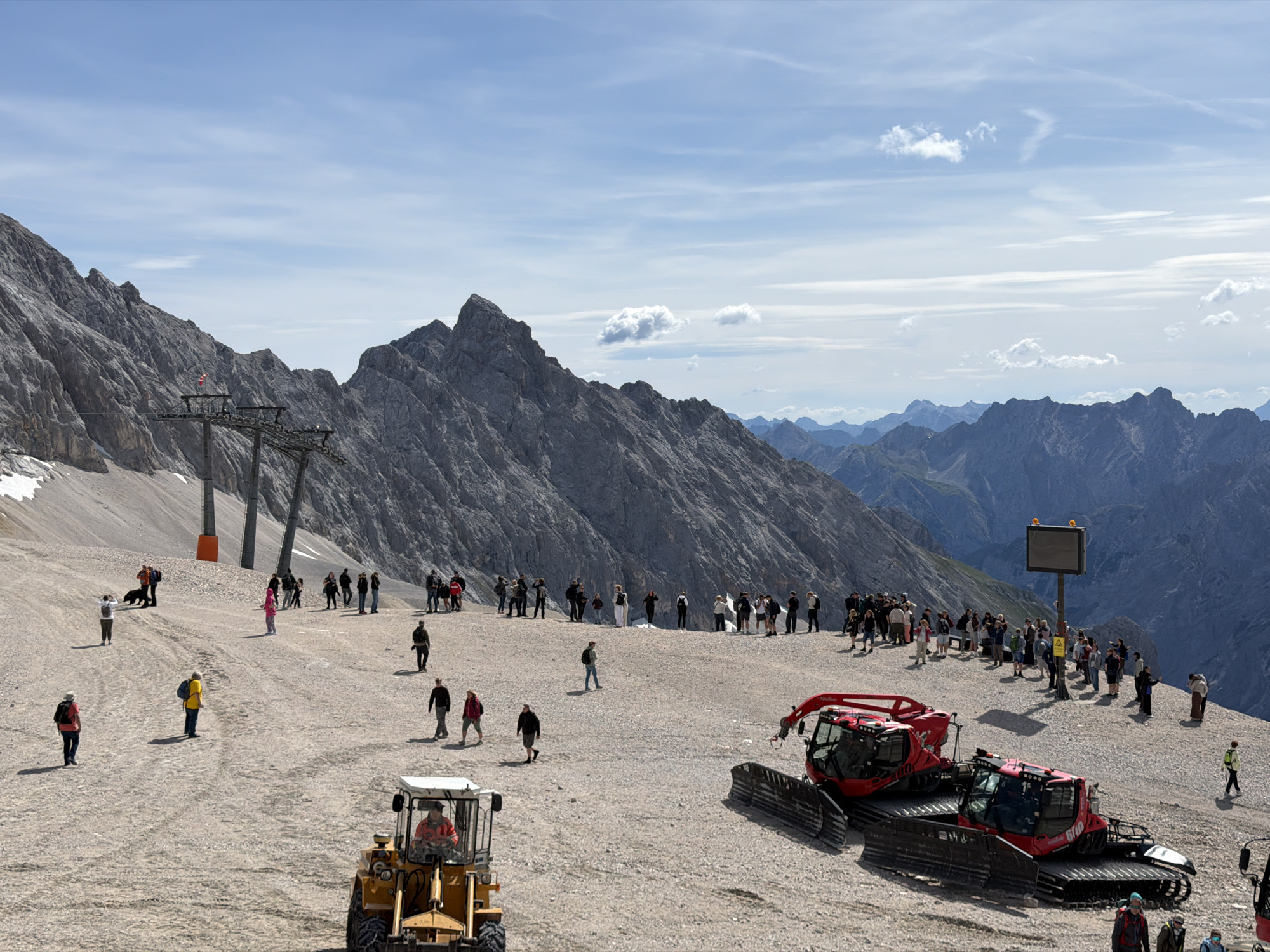 This is a wide-angle, eye-level shot of a gravelly, mountainous landscape, likely taken at the Zugspitze, Germany's highest peak, near Garmisch-Partenkirchen. In the foreground, a yellow bulldozer is parked, facing left, with a person visible in the driver's seat. To the right of the bulldozer, two red snow groomers are parked side-by-side, their tracks facing the viewer.
A group of approximately 30-40 people are scattered across the gravel expanse, some standing and observing, others walking. Most are dressed in casual, outdoor clothing. Two cable car towers rise from the left side of the frame, supporting cables that extend towards the mountain. The mountains themselves are rugged and rocky, with patches of snow visible on the higher slopes. In the background, a chain of distant, hazy blue mountains stretches across the horizon under a sky with scattered white clouds. A digital display board is mounted on a pole in the mid-ground, to the right of the cable car towers. The overall scene depicts a tourist area in a high-altitude mountain environment.