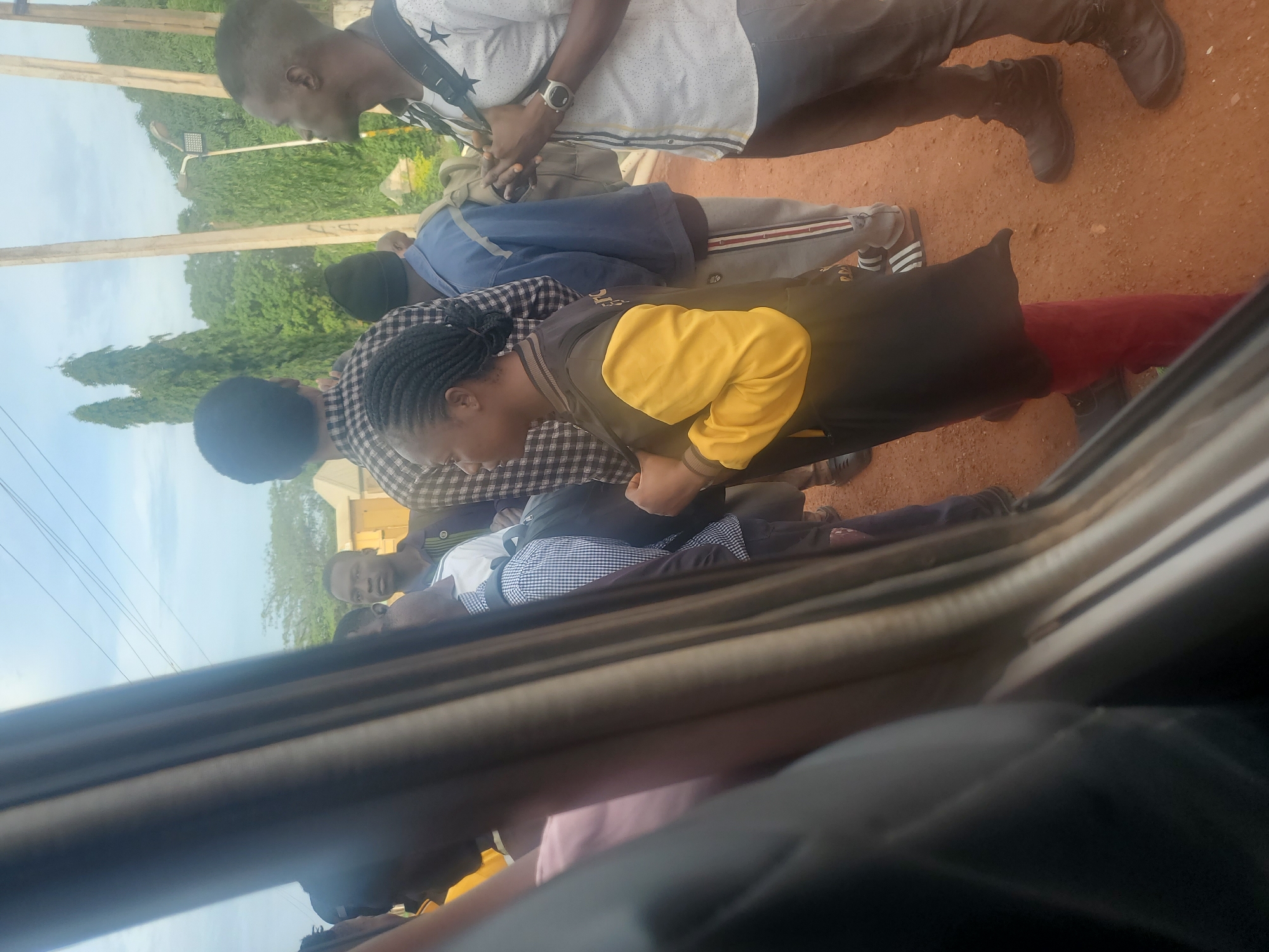In Kakau, Nigeria, a group of individuals stands outdoors, some appearing to be near a vehicle. The scene is outdoors, with a cloudy sky and lush green trees in the background, suggesting a tropical or subtropical climate. The ground is a reddish-brown, likely soil. A utility pole with wires is visible, along with a streetlamp mounted on another pole.
Several people are present, with their clothing indicating a casual or everyday setting. One woman, wearing a yellow and black jacket over red pants, stands prominently in the foreground. To her right, a man in a white shirt with black star patterns and dark pants is visible. His hands are clasped in front of him, and he wears a watch on his left wrist. Another man, wearing a checkered shirt, is seen looking away from the camera. Several other individuals are partially visible, their attire including a blue jacket and a white checkered shirt. The arrangement of people suggests they may be waiting or conversing. The overall impression is a snapshot of a moment in time, capturing a diverse group of people in an outdoor environment.