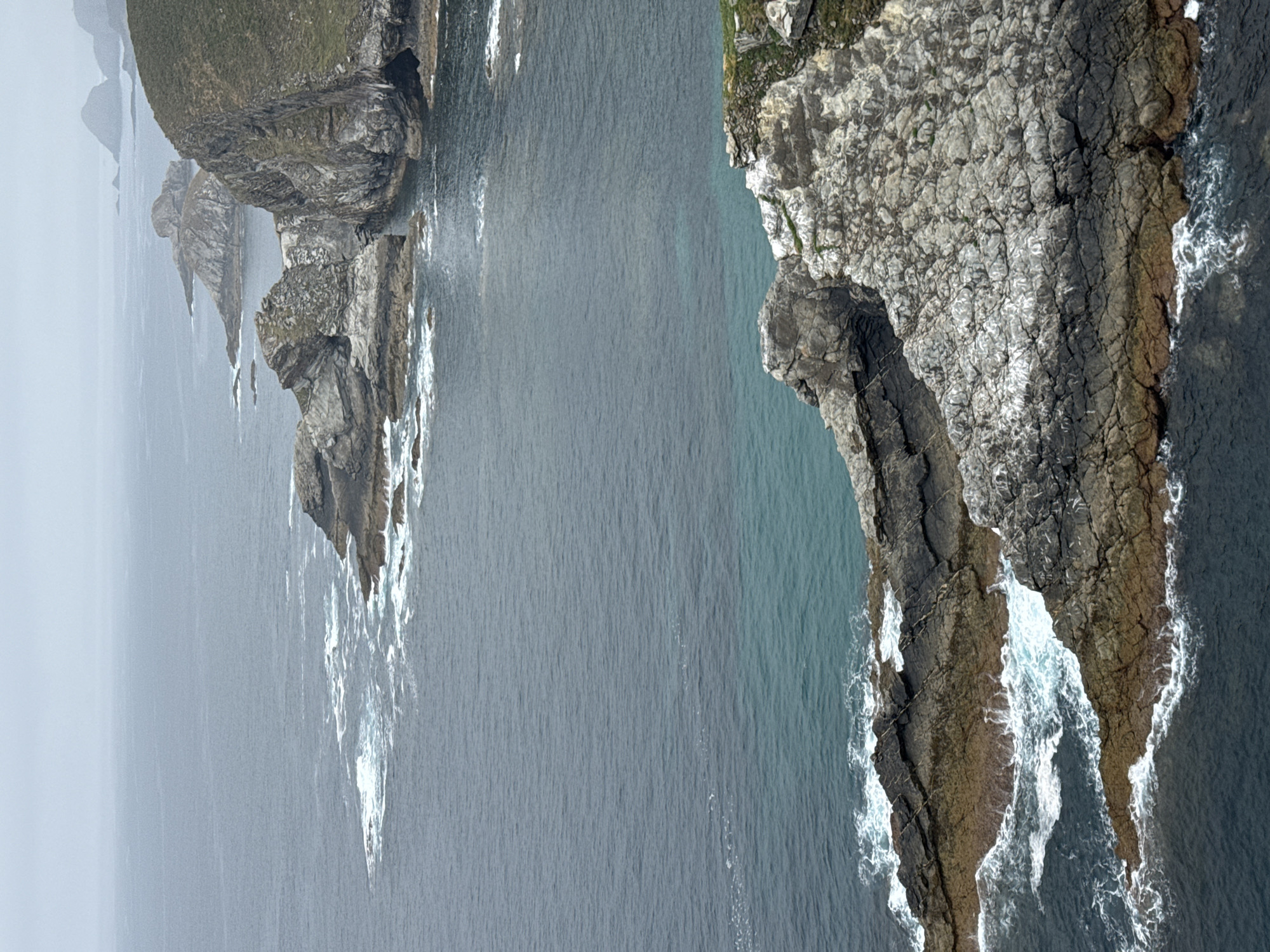The image shows a dramatic coastal landscape with rugged rock formations jutting out into the sea. The water is a deep, dark blue, transitioning to a lighter turquoise in shallower areas. Waves crash against the rocks, creating white foam. The cliffs are steep and covered in patches of green vegetation. In the distance, more rocky outcrops and islands are visible under a hazy, overcast sky. This scene is characteristic of the wild coastline near Cudillero, Spain, a region known for its dramatic cliffs and sea stacks. There are no people or man-made structures visible in the image. The environment is wild and untouched, dominated by the raw power of nature.