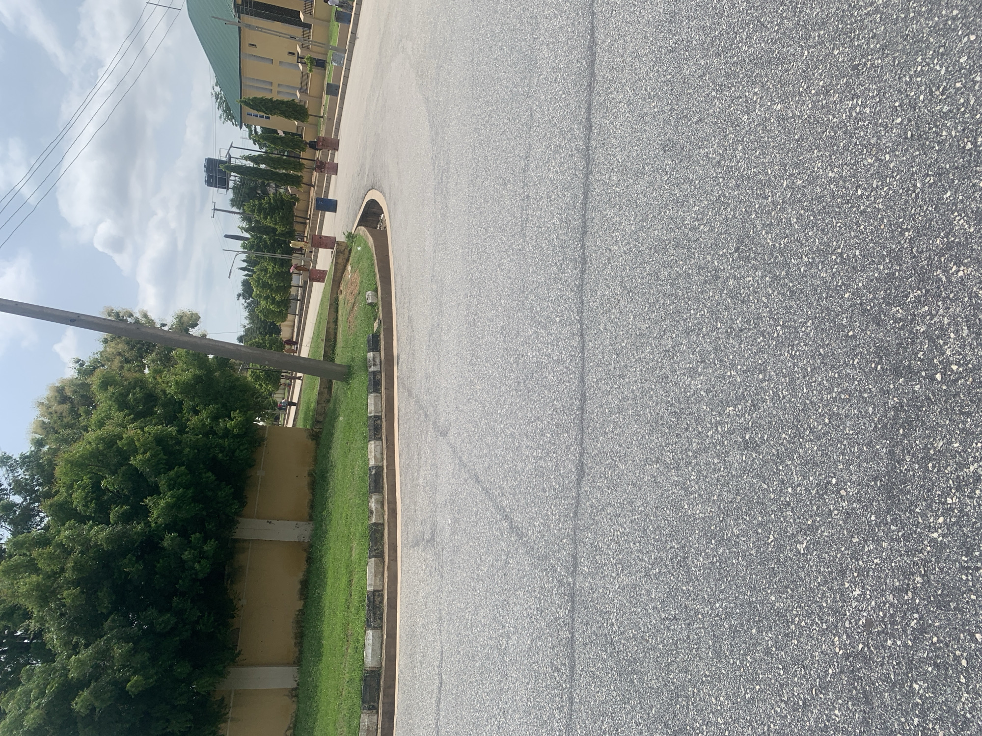 The image captures a street scene in Jalingo, Nigeria, likely in a well-maintained area with a building and extensive landscaping. A paved road curves through the frame, with a black and white striped curb bordering a strip of vibrant green grass. Beyond the grass, a section of a light yellow wall is visible, partially obscured by lush, dark green trees. In the background, a modern building with a green roof and yellow facade stands prominently. Several people are present, walking along the sidewalk. A tall wooden pole, possibly for utility lines, stands in the foreground. The sky is a mix of blue and white clouds. The overall impression is of a clean, orderly urban environment.
