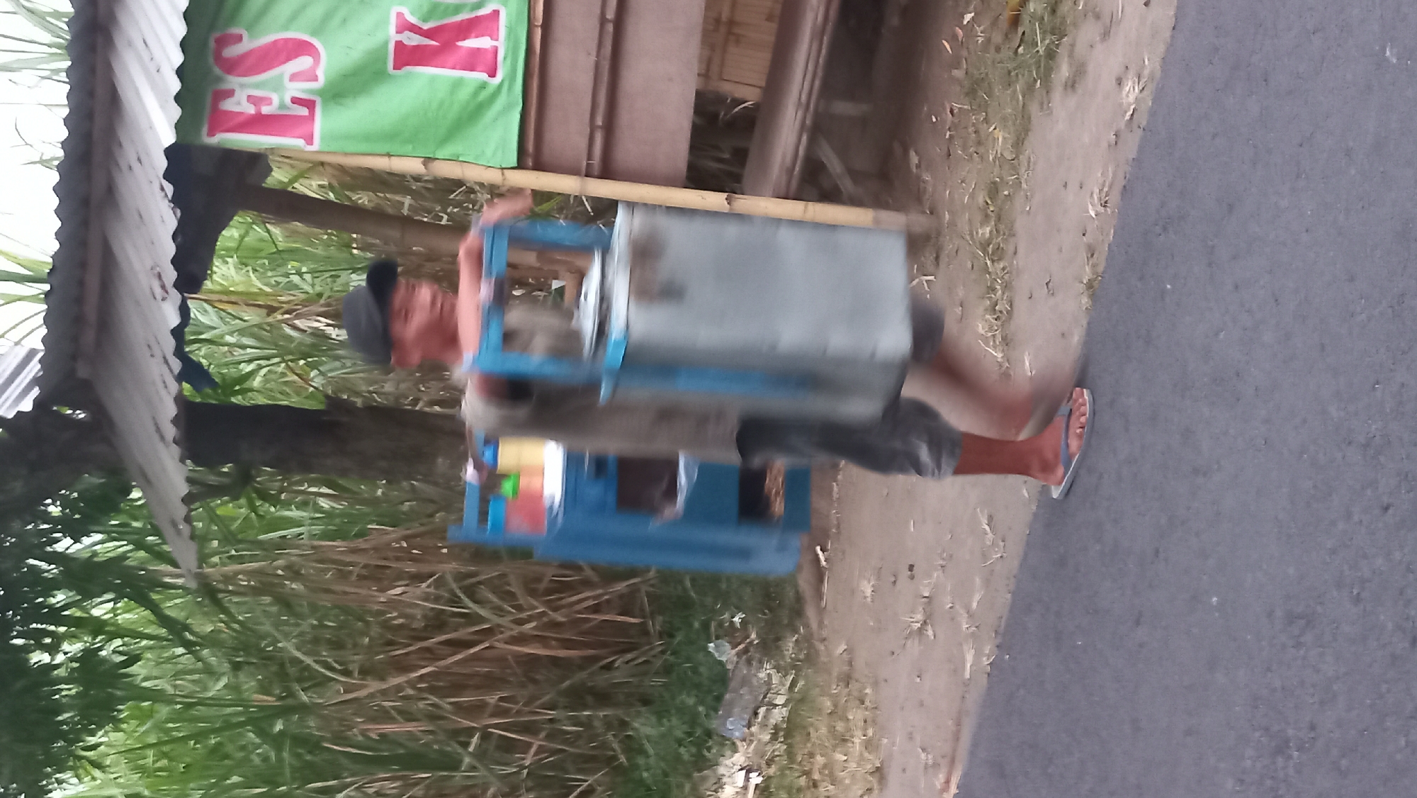 A young male, wearing a black cap, a grey t-shirt, and grey shorts, is walking along a dirt road next to an asphalt road. He is carrying a blue ice chest with a silver metal container on top, likely for selling ice. In the background, there is a green banner with red lettering that says "ES K", a small wooden structure with a corrugated metal roof, and tall green plants that appear to be sugarcane. The scene is likely in a rural area of Pare, Indonesia, given the presence of sugarcane and the typical roadside vendors. The lighting suggests it is daytime, and the overall atmosphere is casual and everyday.