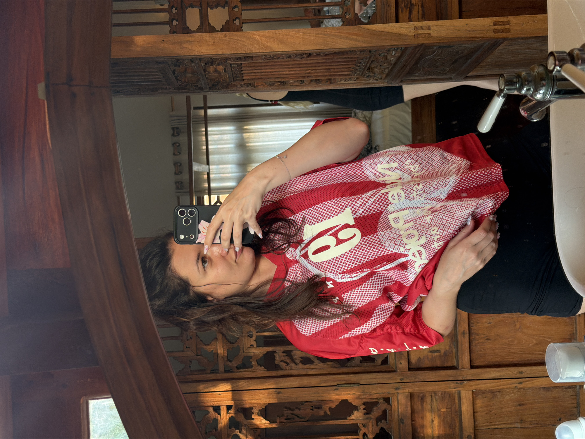 A female is observed taking a mirror selfie in an interior space consistent with North Kuta, Indonesia. The individual, appearing to be in her 20s or 30s, has long, dark brown hair pulled back in a half-ponytail. She is wearing a red and white striped athletic-style shirt with a large white "9" and other white text patterns, along with black shorts. A delicate silver bracelet with small charms adorns her left wrist. She holds a black smartphone with a white and pink floral-patterned case in her right hand, obscuring part of her face. Her left hand rests near her hip.
The foreground features a white ceramic sink basin with a chrome faucet and white ceramic handles. To the left of the sink, two containers are visible: a white plastic bottle with a blue top and a clear cylindrical container with a white lid.
The background, reflected in the mirror, reveals an intricately carved dark wooden environment, characteristic of traditional Indonesian design. This includes ornate wooden mirror frames, doors, wall panels, and structural beams. To the immediate left of the primary subject, an exterior window allows natural light to enter. Further back, past a carved wooden partition, a room is visible containing a bed with light-colored bedding and a sheer light-colored curtain covering a window. A second female figure, partially reflected, with blonde or light brown hair and wearing a black garment, is visible standing in the background room.