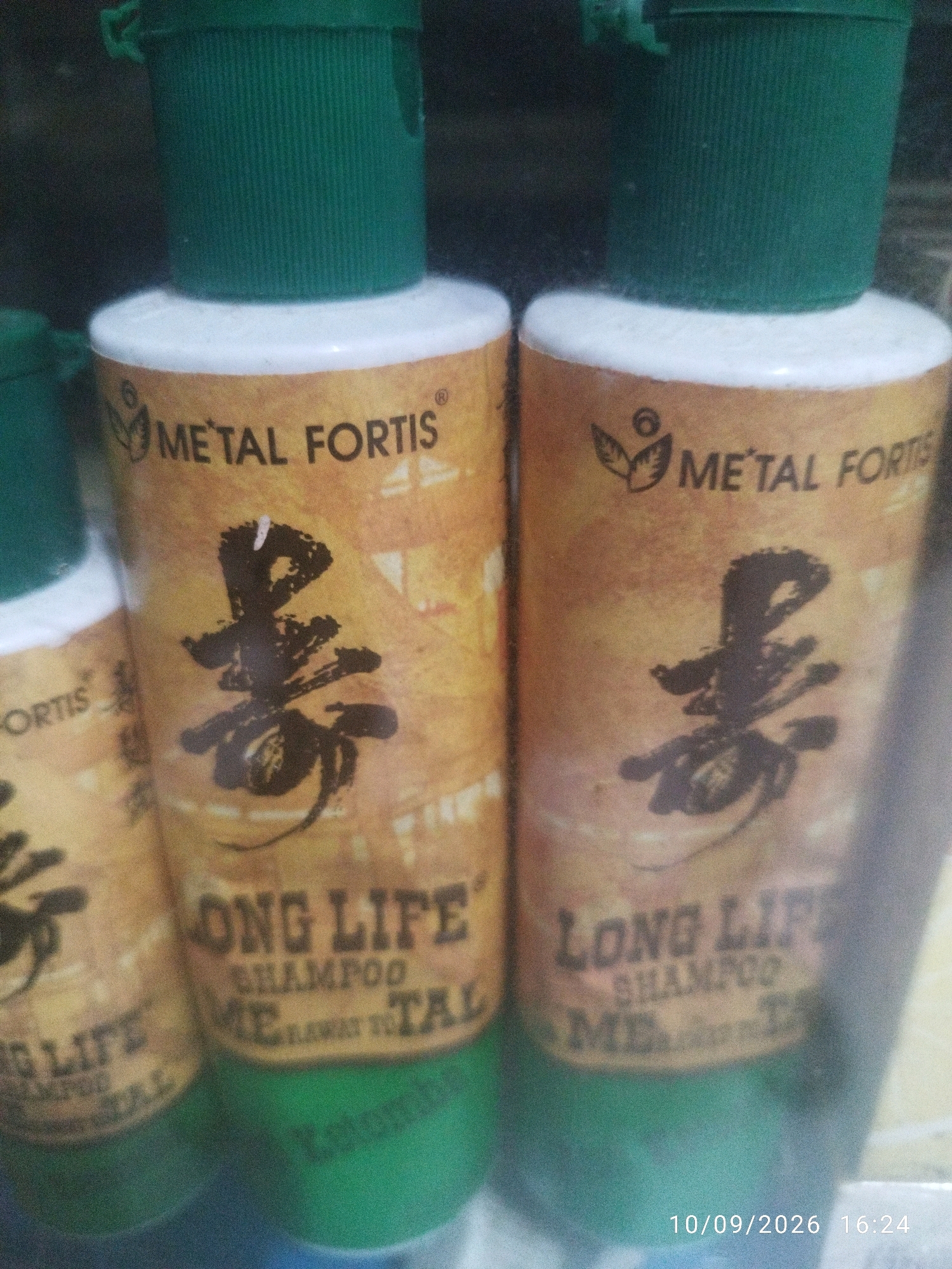The image shows two green-capped bottles of "METAL FORTIS LONG LIFE SHAMPOO" arranged next to each other. The labels on the bottles are predominantly yellow and brown with black text and graphics. A large, stylized black character, possibly Chinese or Japanese, is visible on each label. Below the main text, there is smaller text that is difficult to read, but appears to include "ME" and "TAL". The bottles appear to be plastic and are partially obscured by a green surface at the bottom of the image, which could be a shelf or table. A timestamp at the bottom right of the image reads "10/09/2026 16:24". The lighting is dim, and the image is slightly out of focus. There are no people or any indication of the city of Mojokerto, Indonesia in the image.