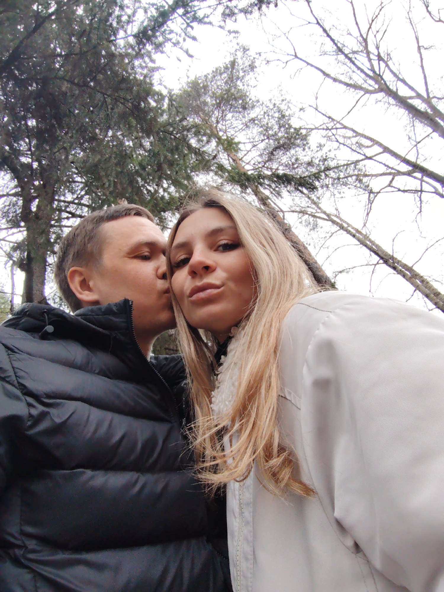 A man kisses a woman in an outdoor setting. The man, wearing a black puffer jacket, kisses the woman on the cheek. The woman has long blonde hair and wears a light-colored jacket with a fluffy collar. Trees with bare branches and evergreen trees are visible in the background. The image was taken in an outdoor setting in Chaikovsky, Russia.