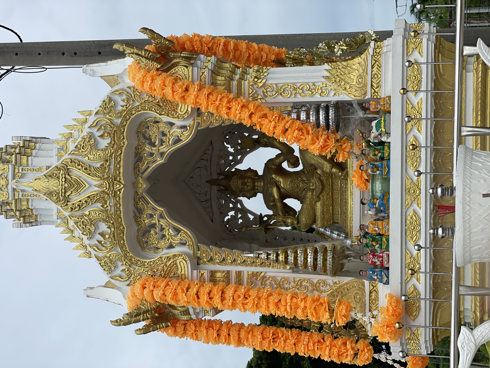 This is a full frontal, medium shot of a golden statue of a deity seated within a white and gold shrine, likely a spirit house, located outdoors in Pattaya City, Thailand. The shrine is adorned with orange marigold garlands on either side, and intricate gold decorative carvings. Beneath the main deity, a row of smaller figures, each in colorful attire, are seated on a ledge. The shrine is situated outdoors, with a cloudy sky overhead and a utility pole visible on the right side of the frame. The shrine appears to be elevated on a white base, with metal railings in the foreground, suggesting it is in a public area. The overall impression is one of religious devotion and cultural display.