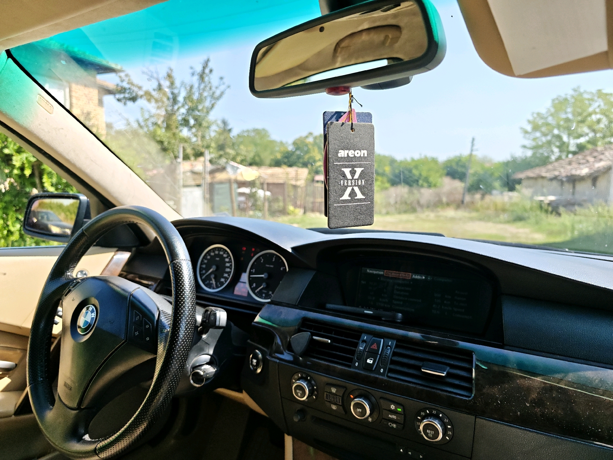 The image is taken from inside a car, looking out through the windshield. The dashboard is visible, with a steering wheel on the left and a navigation screen in the center. A car air freshener hangs from the rearview mirror. Outside, there are trees and buildings, suggesting a rural or suburban setting. The scene appears to be in Karamanite, Bulgaria, judging by the car's interior and the surrounding environment.