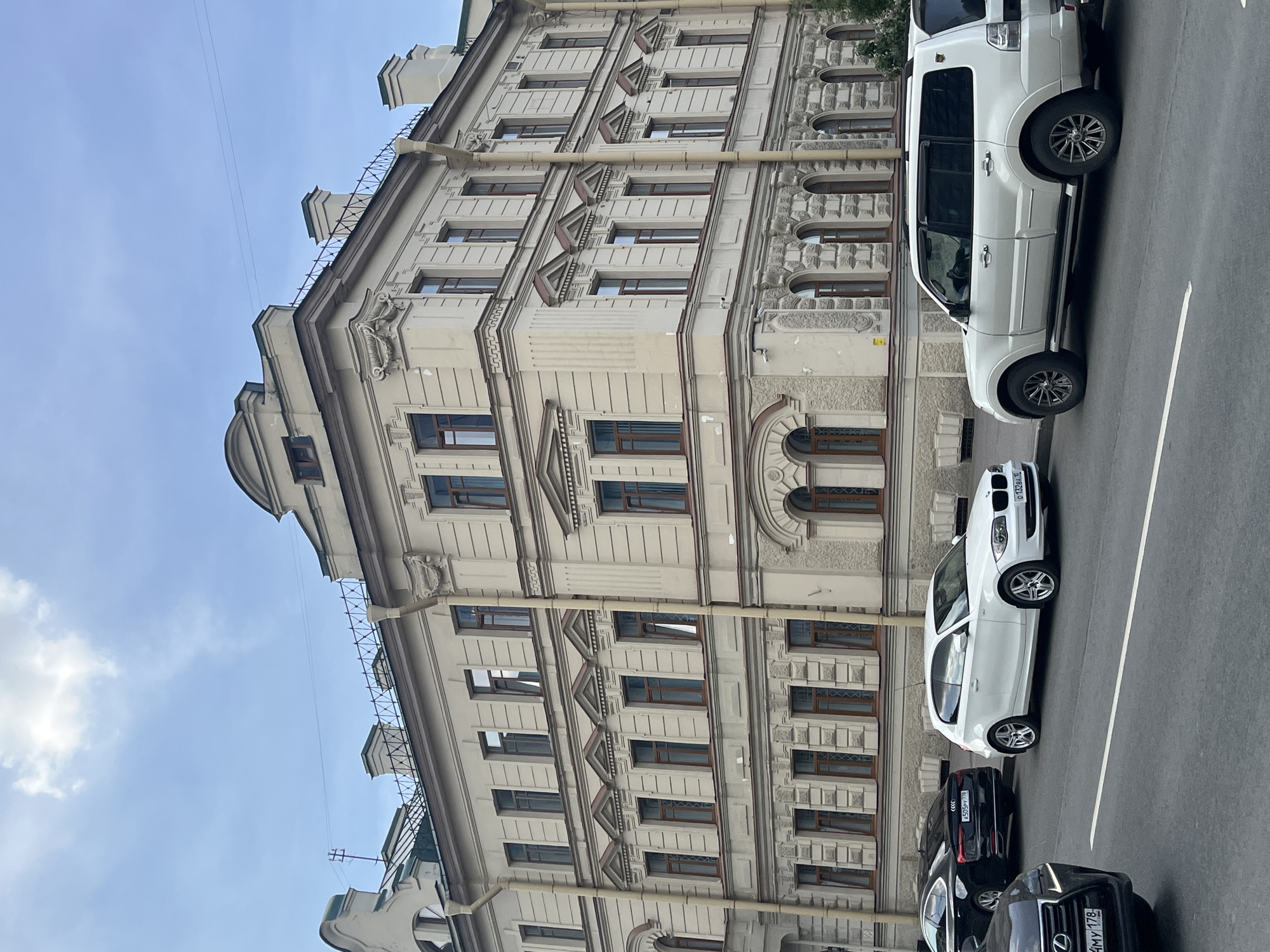 The image shows a street scene in Saint Petersburg, Russia, with a large, ornate, pale yellow building dominating the background. The building features multiple stories with numerous windows, decorative cornices, and architectural detailing. On the street in front of the building, several cars are parked and visible. These include a black Audi sedan, a white BMW hatchback, and a white SUV. The sky is partly cloudy. The overall impression is of a classic urban street in a historic European city.