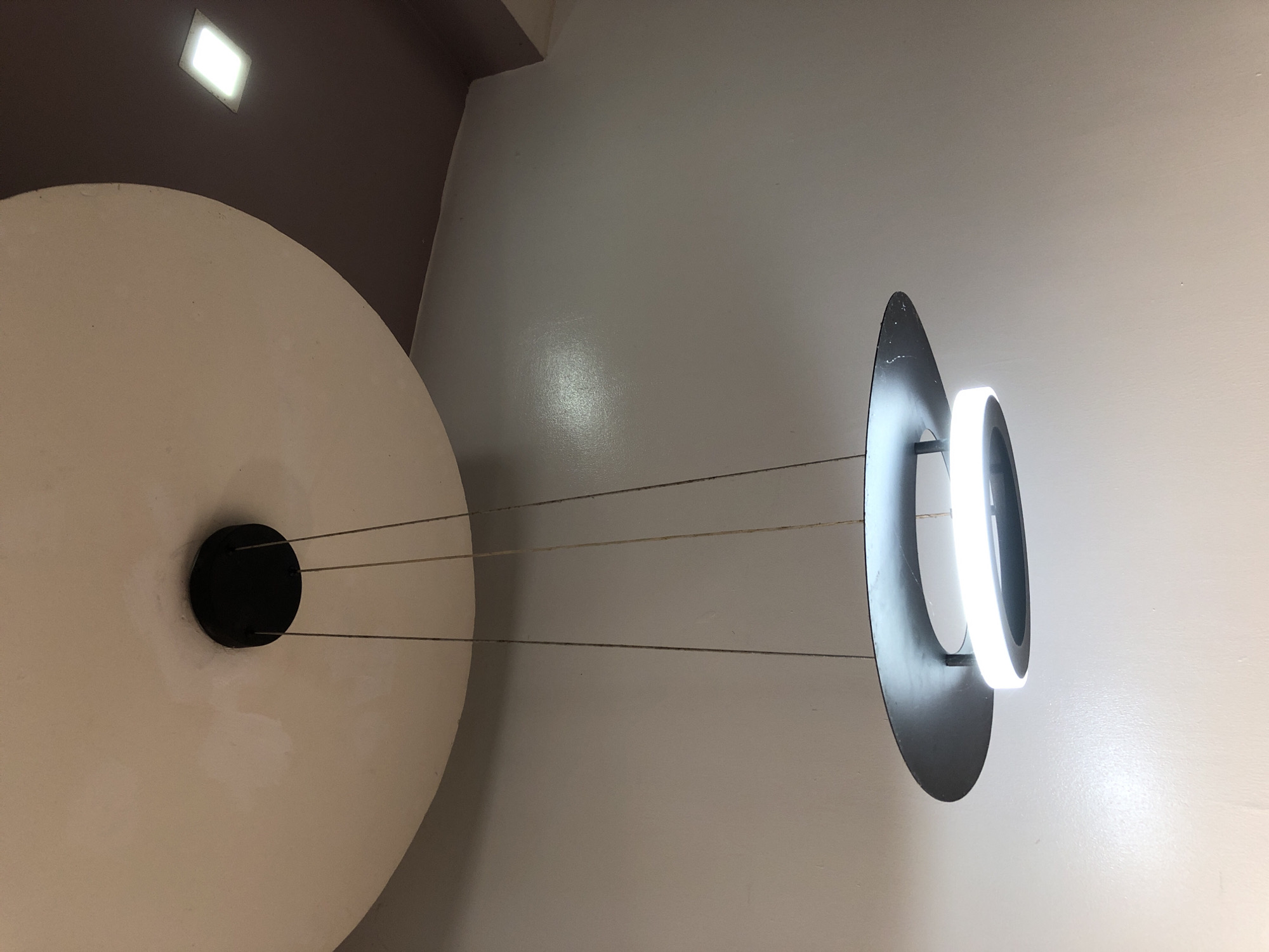 The image displays an indoor scene featuring modern architectural and lighting elements, captured from a low angle looking upwards.
The primary subjects are a large, flat, circular white or off-white object in the lower-left, and a distinctive hanging light fixture towards the right. The white circular object has a smaller, black circular detail at its center, from which four thin, taut wires extend horizontally across the frame. These wires connect to the hanging light fixture.
The light fixture consists of a larger, irregularly shaped, dark (possibly black or dark gray) flat panel or disk, from which a smaller, glowing white ring is suspended. The ring emits bright, cool white light, creating a strong contrast against the darker panel and illuminating the surrounding wall.
The background primarily consists of a light gray or off-white wall/ceiling, with a section of darker brown/purple ceiling visible in the upper-left corner. This darker ceiling section contains a square, recessed light fixture that also emits a bright white light. The overall impression is one of a contemporary interior design, possibly an art installation or a uniquely decorated space like a cafe or lobby.
There is no discernible activity or event taking place, and no people are visible. The scene is static, focusing on the interplay of geometric shapes, light, and texture. The artificial lighting suggests an indoor setting where natural light may be limited or supplemented. No visible text is present in the image.