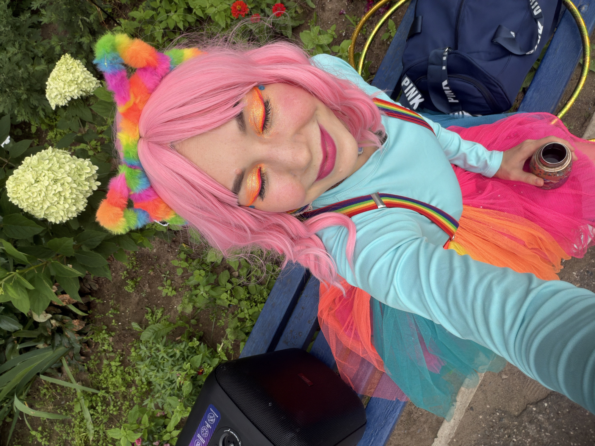 A young woman with bright pink hair and rainbow-colored cat ears is taking a selfie outdoors, sitting on a blue park bench. She is wearing a light blue long-sleeved shirt, rainbow suspenders, and a colorful tutu skirt in shades of pink, orange, and teal. Her makeup is vibrant, with orange and glitter eyeshadow. She is holding a metallic cylindrical object in her right hand. To her left is a black portable speaker. To her right, on the bench and on the ground, are a navy blue duffel bag with "PINK" written on it, and a golden hula hoop. The background shows greenery, including some flowering plants, suggesting a park or garden setting. There are no specific identifiable landmarks that would place this scene in the city of Chaikovsky, Russia.