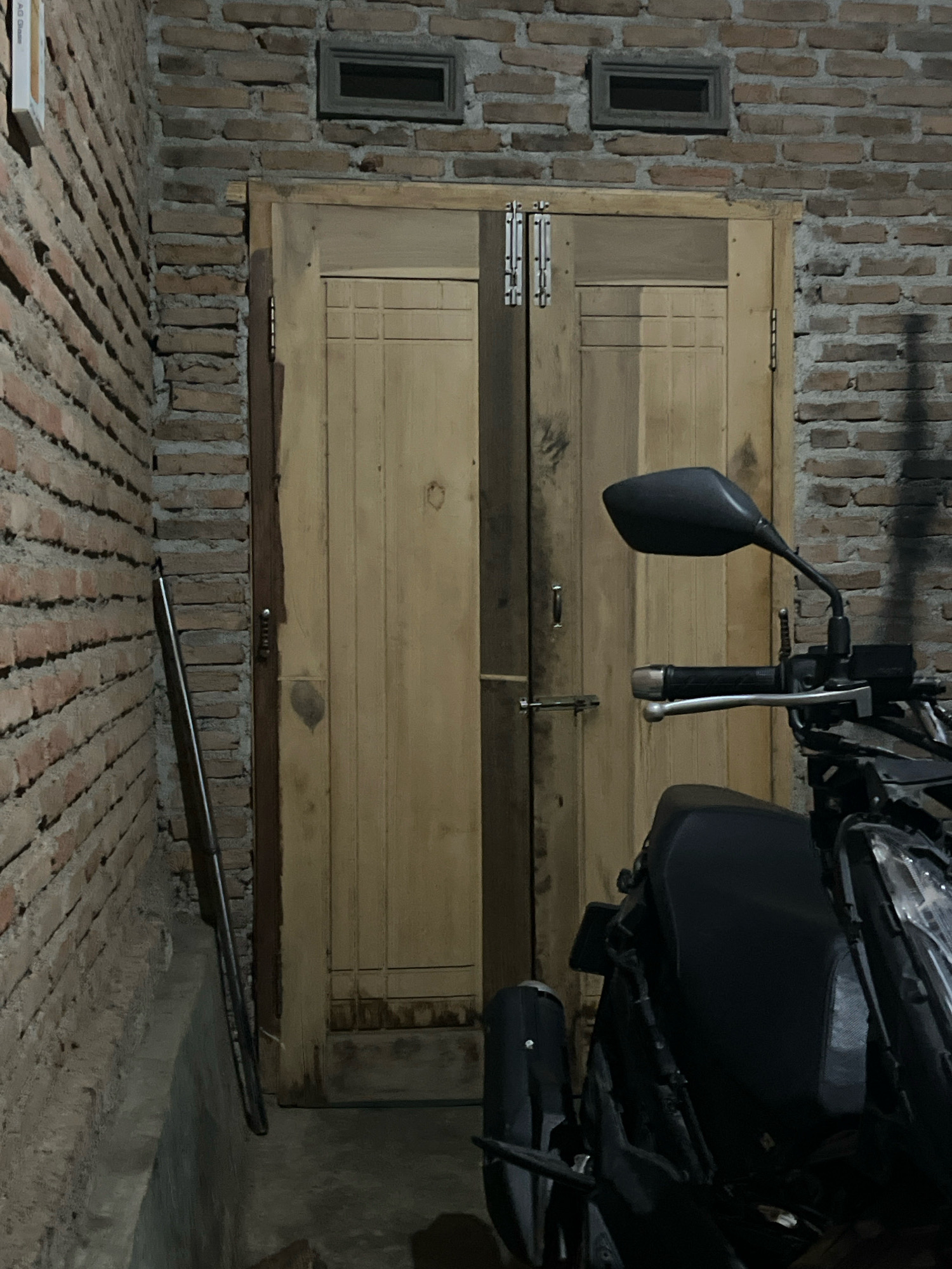 The image depicts a scene at night in Kebumen, Indonesia. The primary focus is a weathered wooden double door set within a brick wall. The doors appear to be slightly ajar, revealing a dark interior. To the left of the door, a long metal pole or pipe leans against the brick wall. In the foreground, the front section of a dark-colored motorcycle is visible, with its handlebars and mirrors prominently featured. The overall environment suggests an outdoor or semi-enclosed space, possibly a driveway or a yard, given the presence of the motorcycle and the brick construction. The lighting is dim, casting shadows and highlighting the textures of the brick and wood. There are no people visible in the frame.