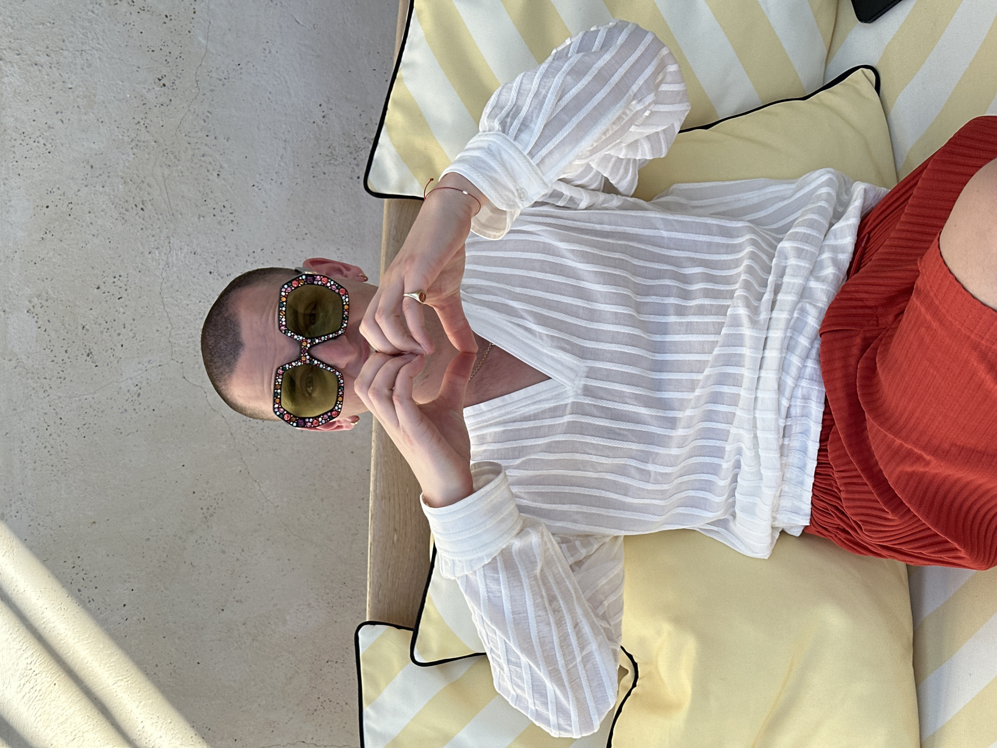 An individual with close-cropped hair is wearing bejeweled sunglasses, a striped white shirt with long sleeves, and red shorts. The person is positioned on striped cushions with black piping, likely part of outdoor seating. The setting appears to be outdoors on a concrete surface. The image could be taken in Sant Josep de sa Talaia, Spain, but there are no readily identifiable landmarks or characteristics confirming the location.