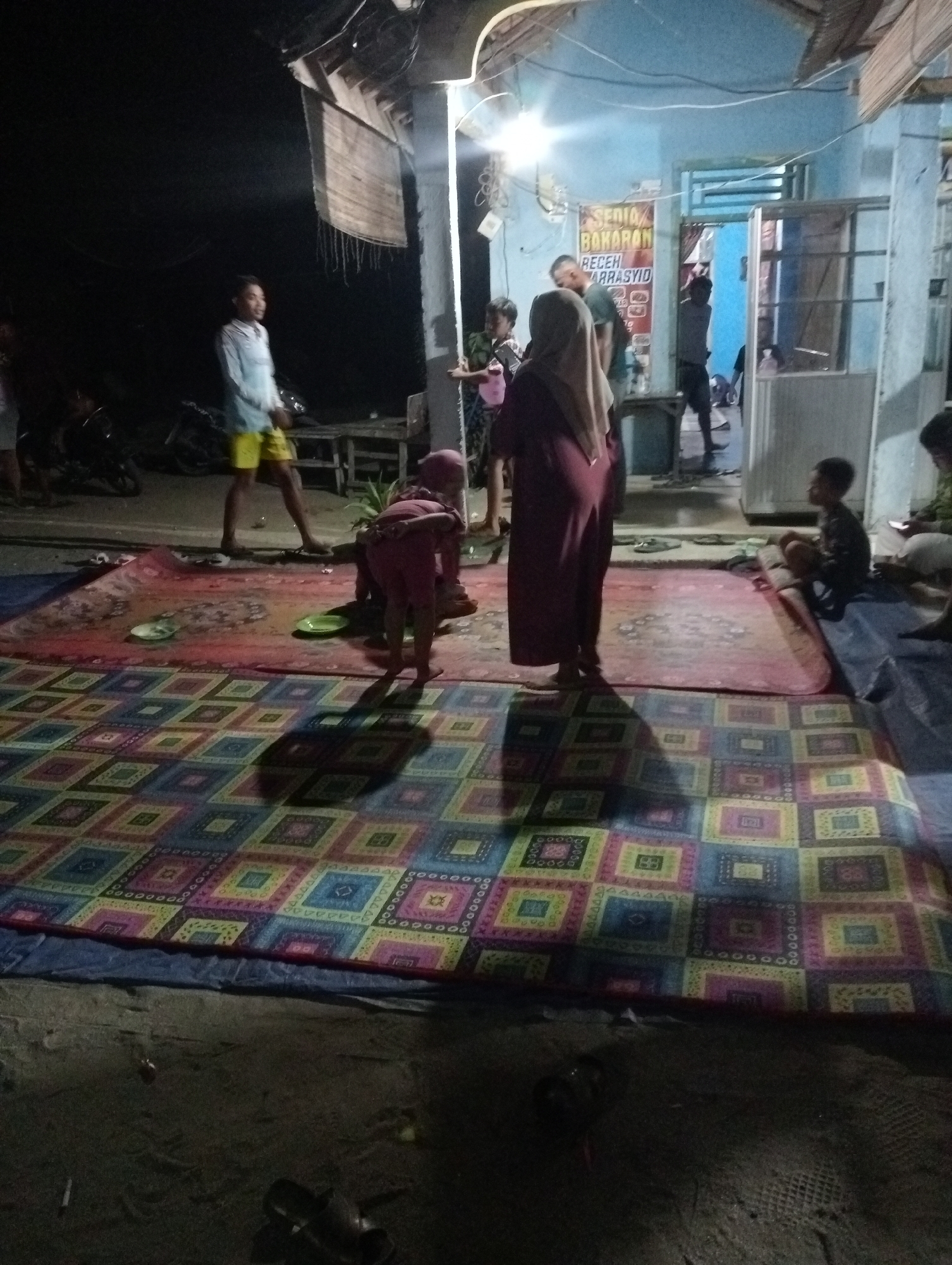 In Rasuan Baru, Indonesia, an outdoor gathering is taking place at night. Several individuals are present, with some sitting and others standing. A young man in a light blue shirt and yellow shorts is standing on the left, facing towards a building with a sign that reads "SEDIA BAKARAN RECEH ARRASYID." Several people are seated on mats and tarps on the ground. A woman in a dark maroon hijab and dress is standing and looking towards the ground. Another person, a child, is sitting on a mat to the right. The ground is covered with several large mats, one with a colorful geometric pattern. The environment appears to be a residential area, with a building in the background and what appears to be a motorcycle parked on the left. The lighting suggests artificial illumination from the building.