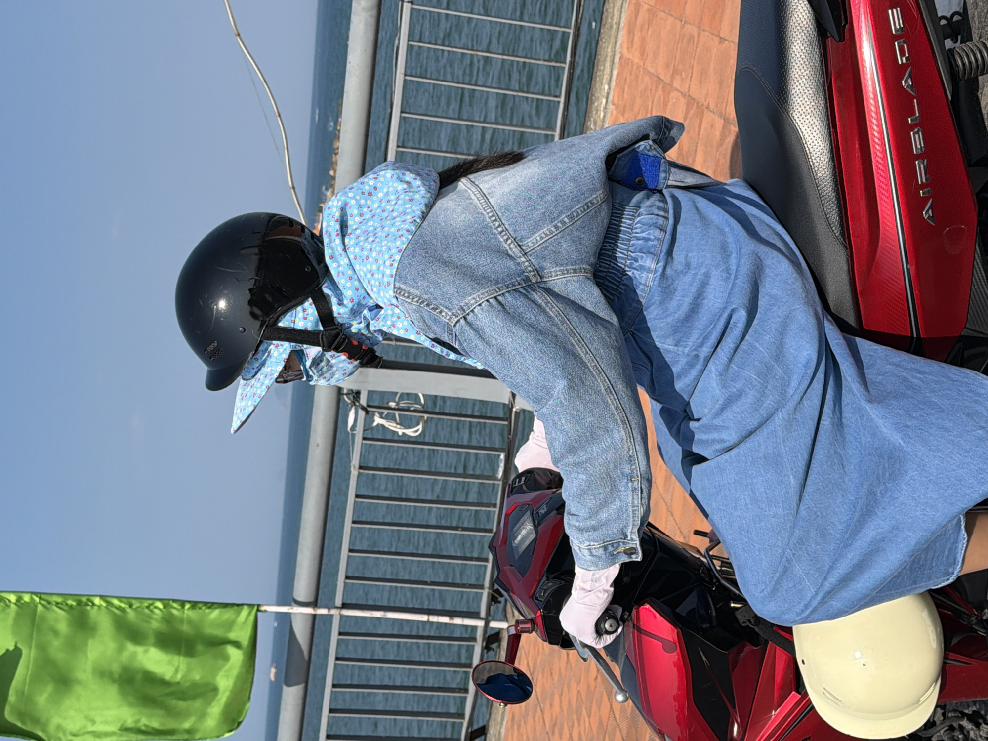 A person wearing a black helmet, a blue patterned scarf over their face, a denim jacket, and blue pants rides a red scooter. The scooter has a black seat and a cream-colored object, possibly another helmet, attached to the front. The person is wearing light pink gloves and is holding the scooter's handlebars. They are positioned on a paved walkway next to a metal railing that overlooks a body of water. In the background, the sky is clear and blue, and a green flag is visible on the left side of the image. The scene appears to be a waterfront area in Nha Trang, Vietnam, likely a promenade or a coastal road.