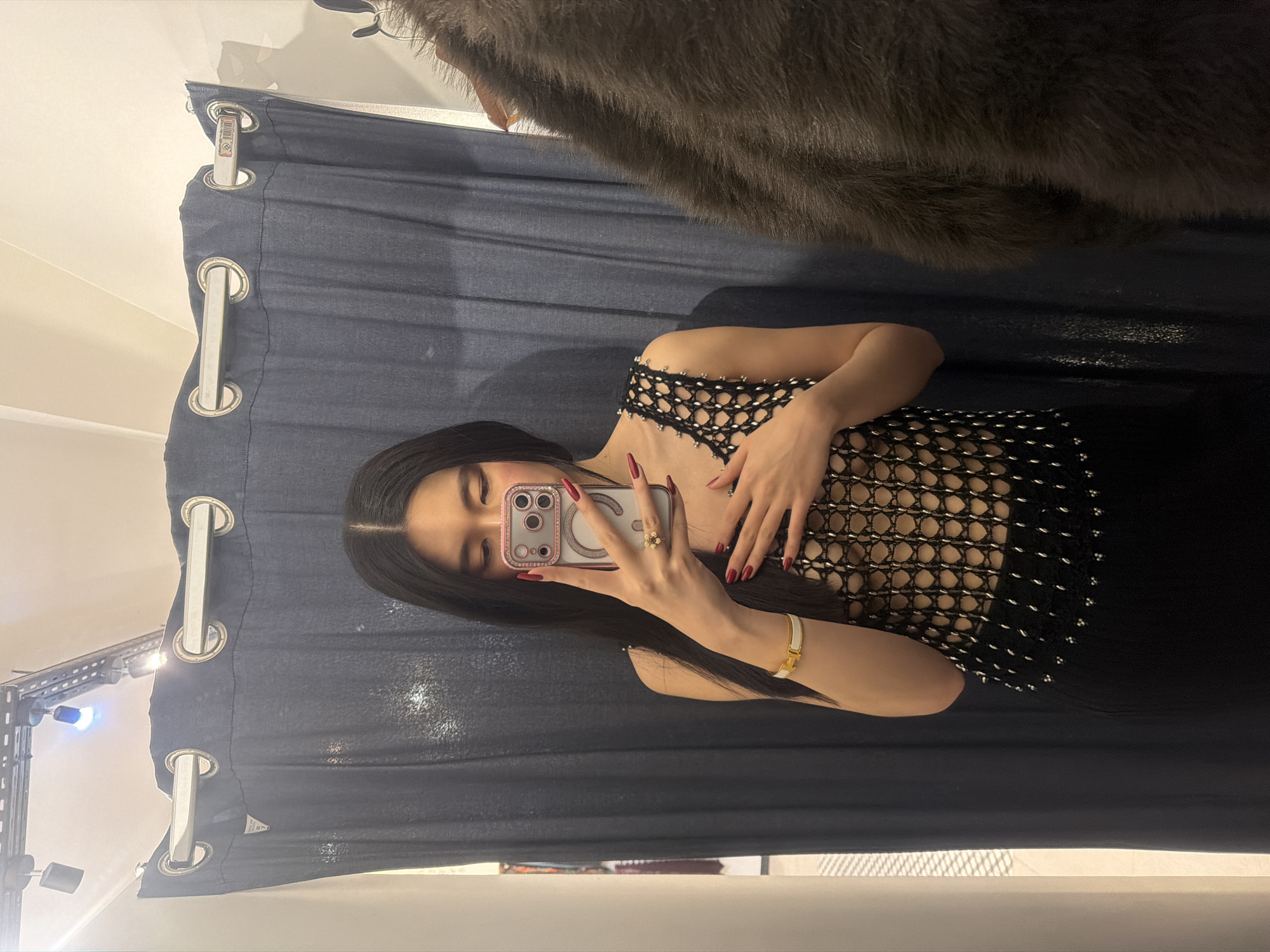A woman with long dark hair takes a selfie with a light pink iPhone. She wears a black, net-like top and a gold bracelet on her right wrist. She is in front of a blue curtain with silver grommets. A light fixture is partially visible on the left side of the frame. A grey, furry object is hanging above the curtain. The image does not contain enough information to identify the location as being in Paris, France.