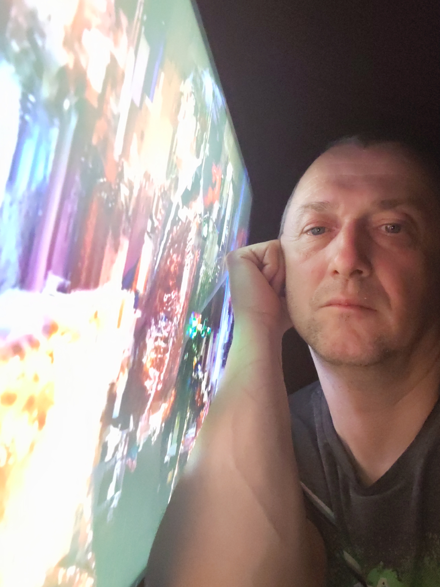 A middle-aged man is positioned in the right of the frame, resting his head on his hand and gazing directly at the viewer. He is wearing a dark gray t-shirt. To the left of the man is a large, brightly illuminated display showing blurred, colorful imagery. The background behind the man appears dark. The location in the city of Volnoga, Russia, is not provided.