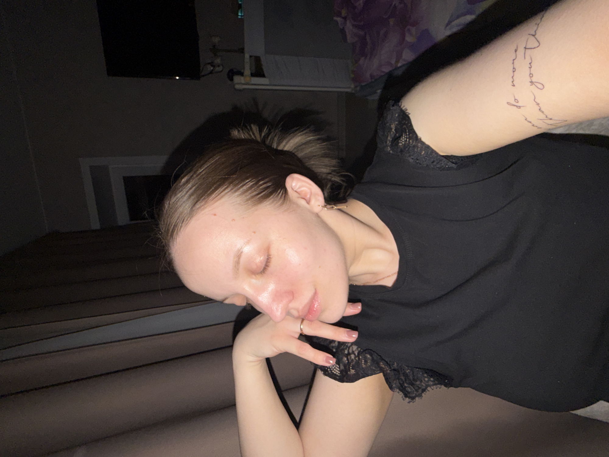 A woman with fair skin and light brown hair is lying on what appears to be a bed. She's wearing a black top with lace trim, and a ring is visible on her finger. A tattoo is on her arm. The background includes what looks like a TV mounted on the wall and a partially visible window. The lighting is dim. There is no information available to determine the location is Reshetnikovo, Russia.