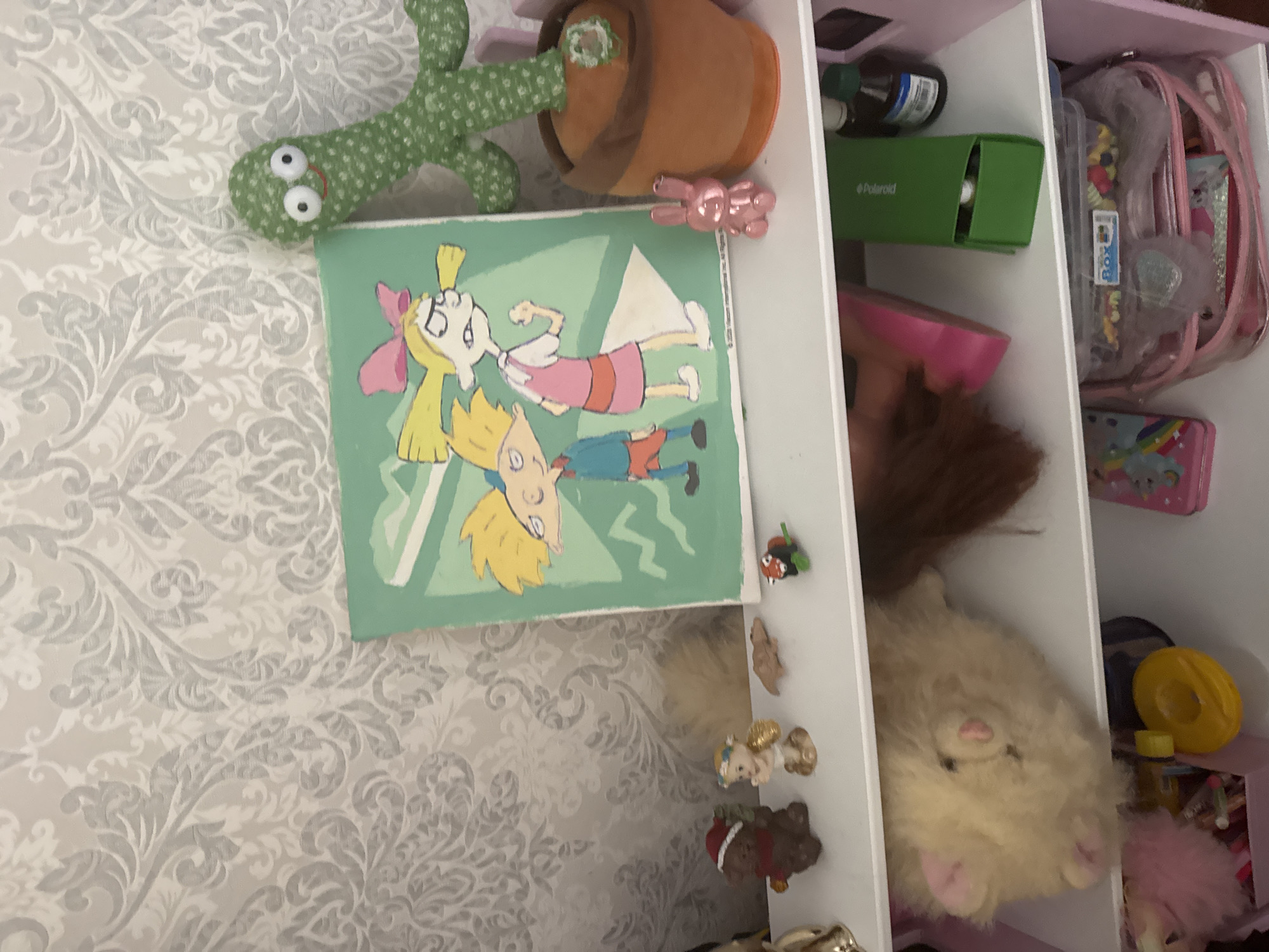 A cluttered shelf unit against a patterned wallpaper displays various decorative items and toys. On the top shelf, a green plush cactus toy sits in a brown pot next to a framed illustration featuring cartoon characters. To the left of the illustration are small figurines of a bear in a Santa hat, an angel, a brown lizard, and a ladybug. The middle shelf holds a fluffy tan stuffed animal, a brown wig, and a pink object. A green Polaroid box and a dark green bottle are also on this shelf. The bottom shelf contains a pink makeup bag filled with items, a clear plastic container with candy, and a pink object with a rainbow design. The environment appears to be an indoor setting, likely a child's room, with a focus on the objects on the shelves.