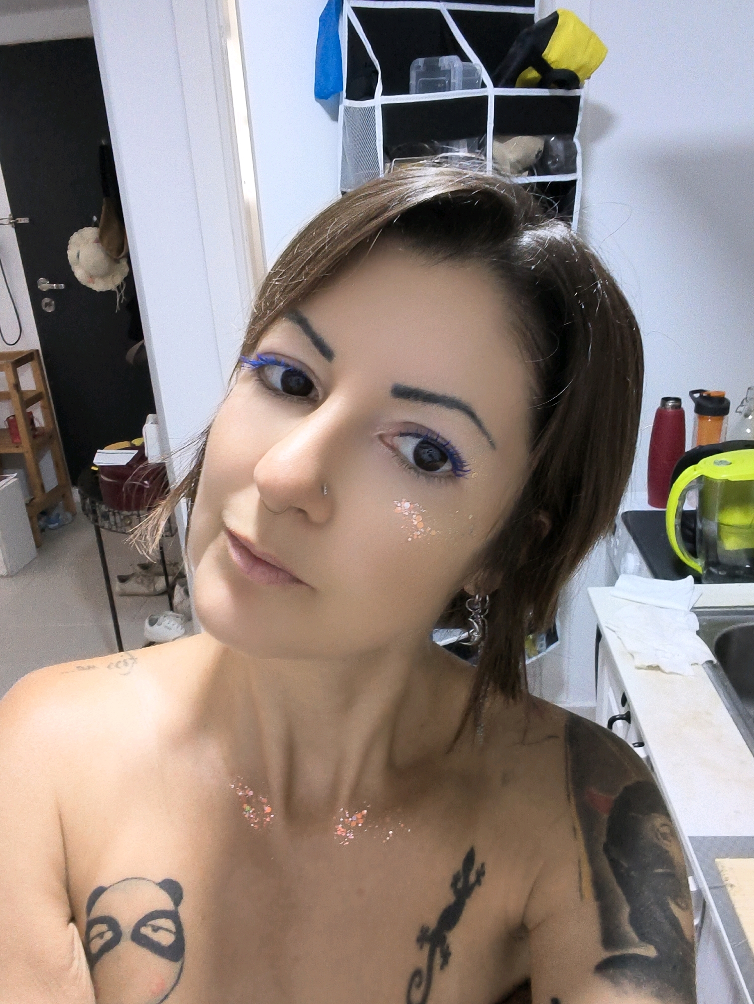 A woman, with brown hair and blue eyeliner, poses for a selfie in an indoor setting. She is wearing glitter on her face and chest, and has tattoos on her shoulders and chest. She is topless. The background shows a doorway, a storage unit with various items, and a kitchen counter with bottles and a sink. The image was taken in the city of Lachish Regional Council, Israel.