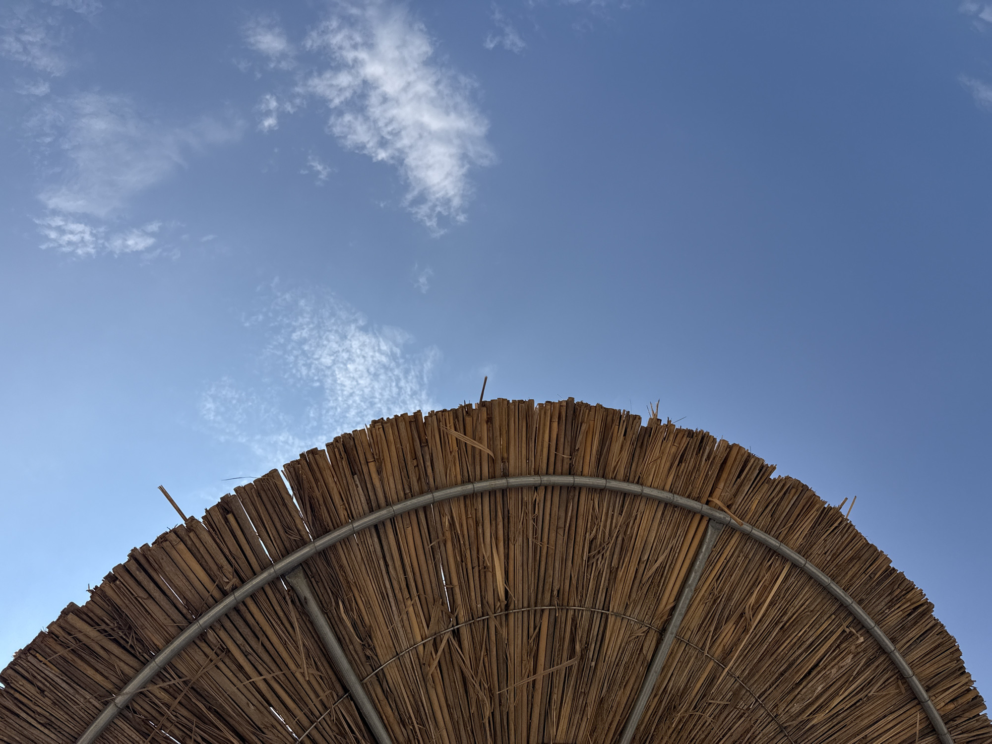 An outdoor, low-angle, close-up, eye-level, full shot depicts the underside of a thatched umbrella against a bright blue sky with sparse white clouds. The umbrella, made of dried reeds, is positioned on the left half of the frame, curving from the bottom left corner upwards and to the right. A metal band encircles the edge of the umbrella, supporting the thatched material. The sky occupies the right side of the image, with delicate, wispy clouds scattered across it. There are no people visible in the image. The environment appears to be a sunny day, likely at a beach or outdoor recreational area. The location is identified as Sithonia Municipal Unit, Greece.