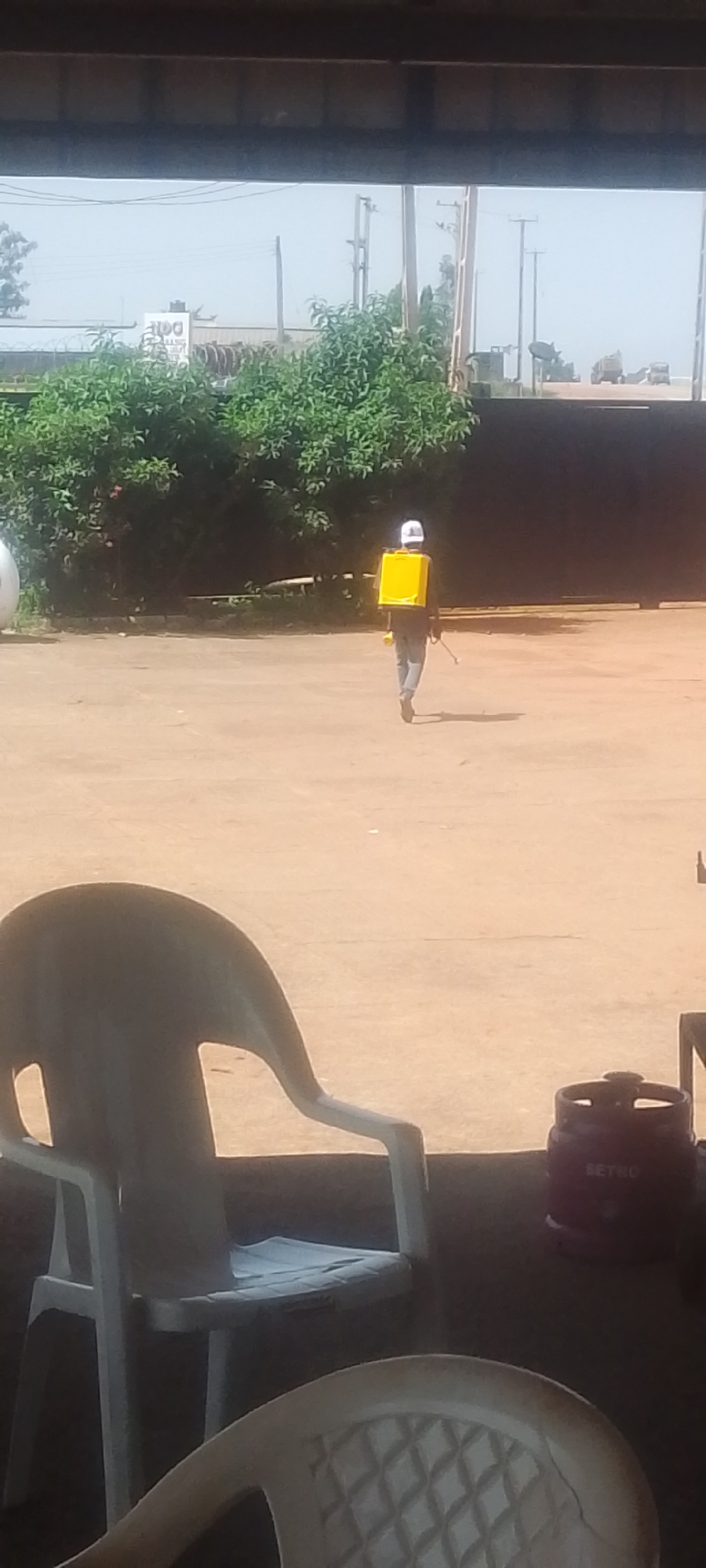 In the city of Jos, Nigeria, a person wearing a white hard hat and a yellow backpack sprayer walks away from the camera in an outdoor, open area with a light brown, dusty ground. The person is wearing a dark shirt and blue jeans. The backpack sprayer is rectangular and yellow, with a hose and wand extending to the person's right. Behind the person is a dense line of green trees and foliage, and further in the distance, a large dark gate or wall. The foreground shows parts of white plastic chairs and a red propane gas cylinder with "BETRO" written on it, suggesting this scene is viewed from a covered area, possibly a patio or veranda. The sky is hazy and bright, indicating daylight. In the background, there are power lines and poles, and a few vehicles are visible on a road beyond the gate.
