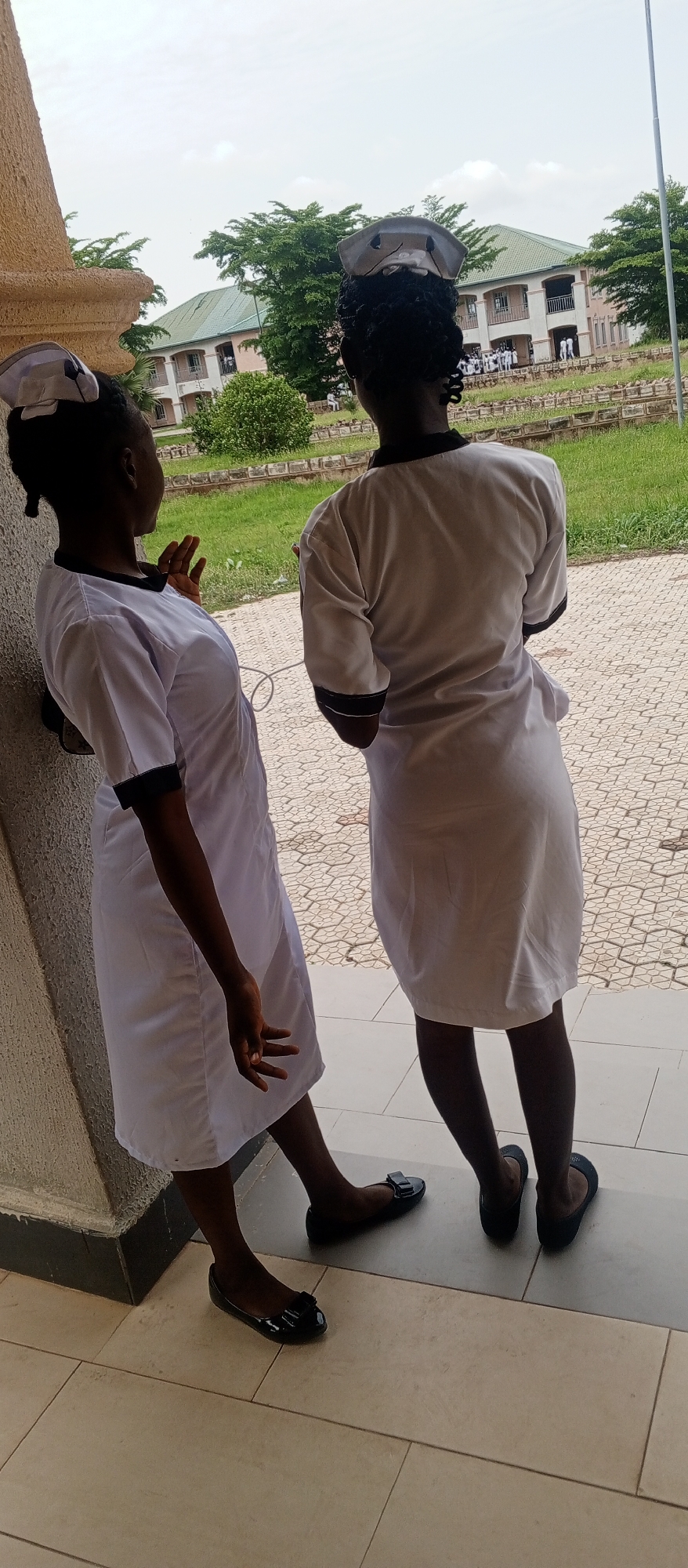 Two individuals, dressed in white nurse uniforms and caps, stand outdoors. The individual on the left is leaning against a textured, light brown pillar and facing away from the camera, with their left arm resting on the pillar and their right hand extended. The individual on the right is standing with their back to the camera, looking towards a building in the background. Both individuals are wearing black ballet flats. The environment appears to be a paved walkway or courtyard, with a building and trees visible in the background. The sky is overcast. The location is described as being in Kuson, Nigeria.