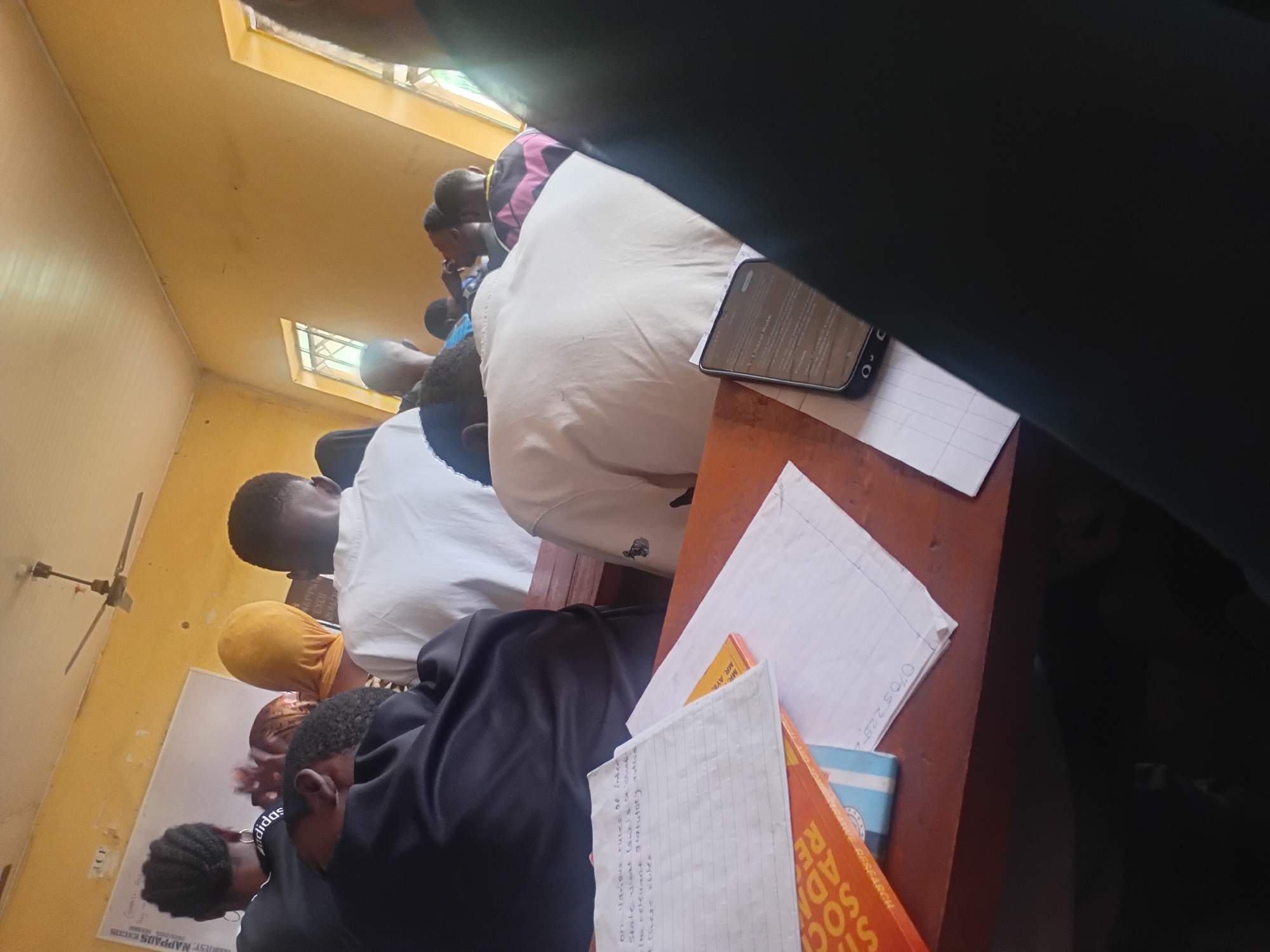 The image depicts an indoor scene within a classroom setting, likely in Ogbogbo, Nigeria, based on contextual information not visually present in the image. The room is characterized by its yellow-painted walls and ceiling, with fluorescent lighting fixtures and a ceiling fan visible. Sunlight streams in through two windows on the right side of the frame, illuminating the space.
Several individuals, primarily young adults or teenagers, are seated at wooden desks, facing towards the front of the room. Most are seen from behind or the side, engaged in what appears to be a lecture or study session. Their attire is casual, with some wearing white shirts, a black top with "Adidas" branding, and a yellow head covering.
On the desks in the foreground, various study materials are arranged. These include notebooks with handwritten notes, a bright orange book with visible text that appears to be related to social research, and a smartphone displaying text, possibly related to legal or academic subjects. The overall atmosphere suggests a typical classroom environment in Nigeria, with students focused on their lessons. The forensic description focuses on the objective details: the color of the walls and ceiling, the presence of furniture and educational materials, and the posture and apparent engagement of the students.