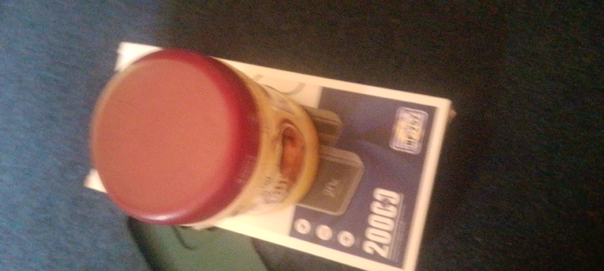 The image shows a close-up of a round, reddish-pink object with a metallic sheen, possibly a container lid, resting on a white and blue box. The box has a pattern of small circles and the number "2000CJ" printed in large, white, stylized lettering. There is also a small rectangular logo with text on the box, partially obscured by the round object. The lighting is dim, suggesting an indoor setting, possibly at night or in a dimly lit room. The background is a dark, textured surface, possibly a table or fabric. The image is slightly tilted and out of focus in some areas, making it difficult to discern finer details. The overall impression is of a product shot or a casual photograph of an item placed on packaging. There are no people or animals visible in the image. Given the location context of Jalingo, Nigeria, the objects could be related to local products or consumer goods, but there are no explicit clues to confirm this.