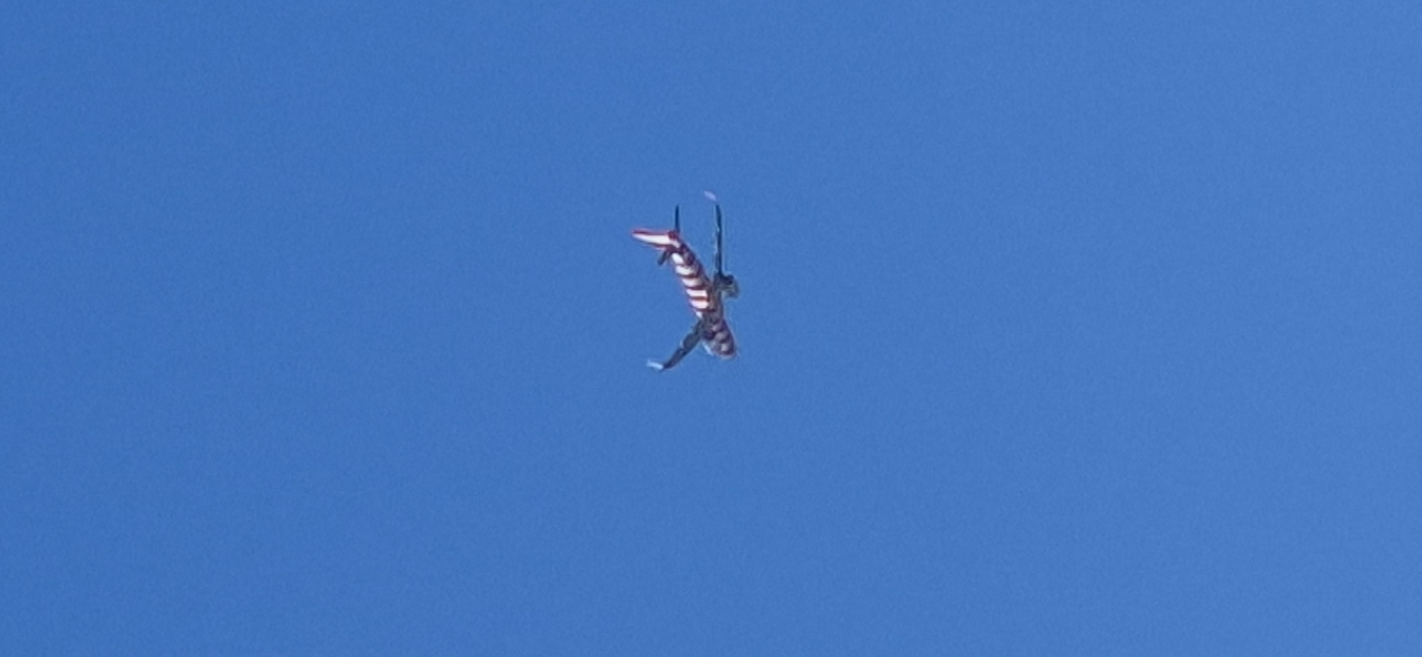 A passenger airplane with a red and white striped livery is seen flying in a clear blue sky. The plane is angled, with its nose pointing slightly upwards, and its wings are extended. The distinctive red and white stripes cover the fuselage and tail of the aircraft. The image captures the plane from below, high in the atmosphere, with no visible ground features or city landmarks, making it impossible to determine its location in Mainz, Germany.