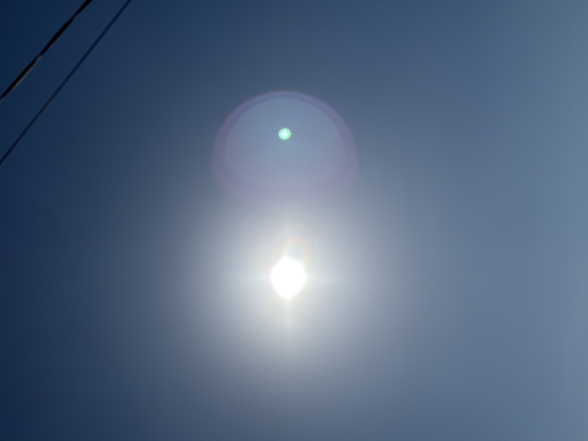 The image shows a bright sun in a clear blue sky, with a subtle lens flare to its right. Two power lines are visible in the upper right corner of the frame, casting faint shadows. The lower portion of the image transitions to a lighter shade, possibly indicating the horizon or atmospheric haze. There are no people or specific landmarks visible to determine a precise location within Chernihiv, Ukraine.