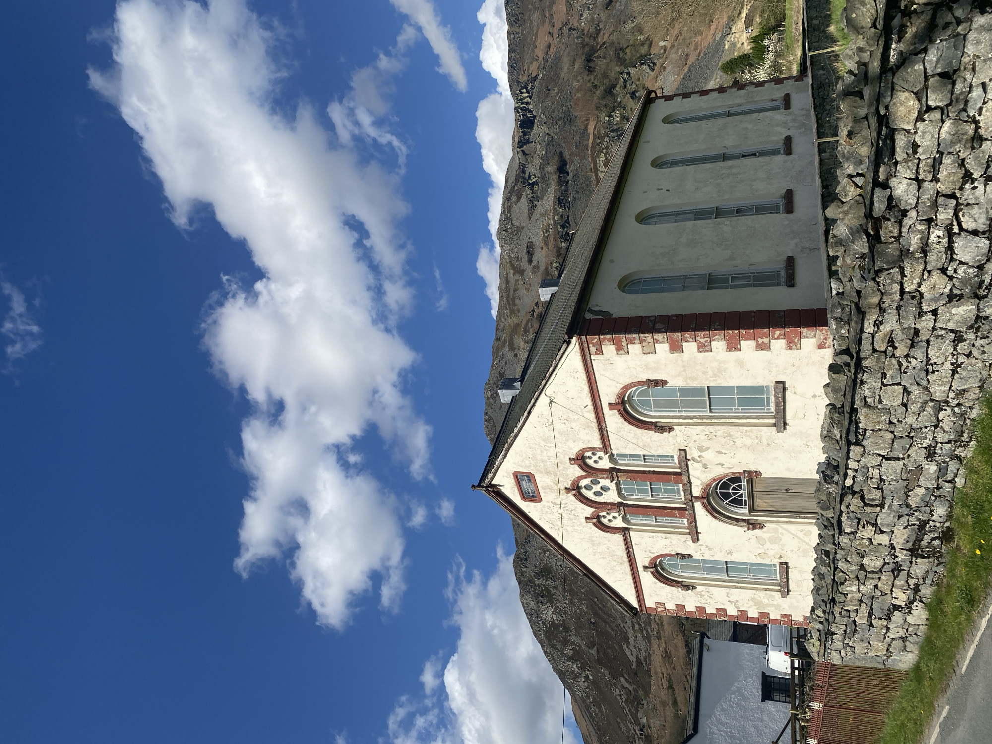 The image shows a building in Oswestry, United Kingdom, next to a rock wall and a mountain. The building is cream-colored with red brick trim and has multiple windows. The sky is blue with white clouds.