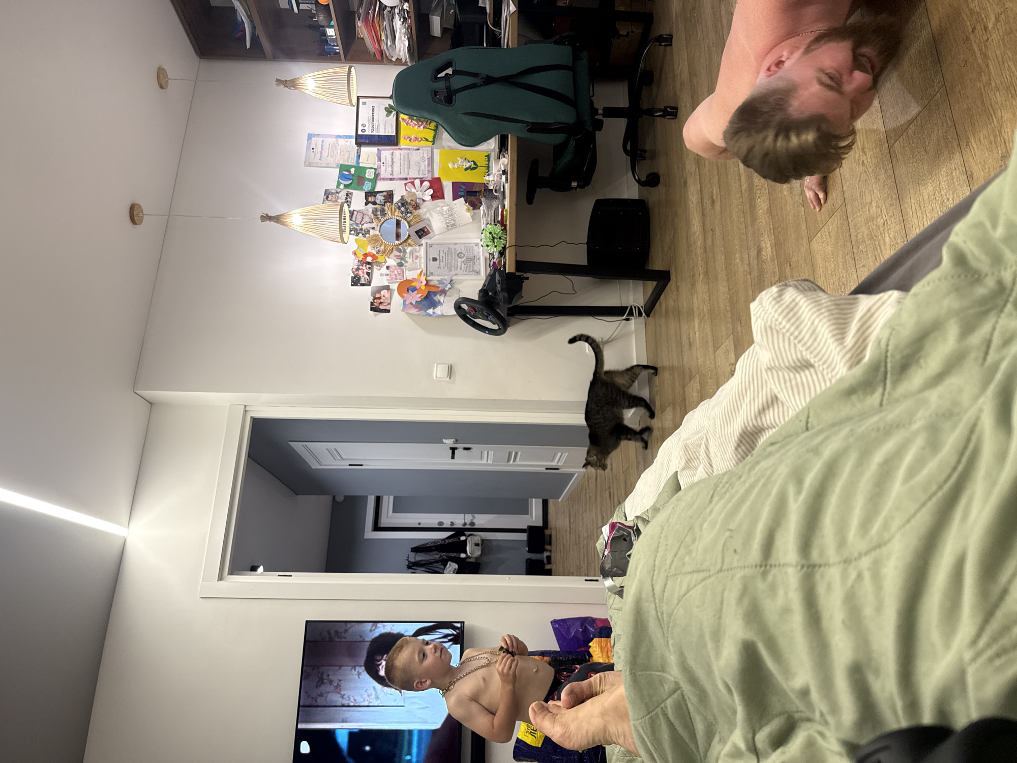 The image depicts an interior scene, likely a living room or bedroom, with an open doorway leading to a hallway. In the foreground, a person's legs are visible, covered by a light green quilted blanket. To the right, a man with a beard and short hair is performing a push-up on a wooden floor. He is shirtless. Further into the room, a young boy, shirtless and wearing a necklace, stands near a television screen displaying a movie or show. Next to him, there are several shopping bags. A tabby cat is walking across the floor between the boy and a desk area. The desk is cluttered with papers, certificates, and various items, including a steering wheel and gaming chair. Two pendant lights hang from the ceiling above the desk. The walls are white, and a linear light fixture is installed on the ceiling. The scene suggests a casual home environment, possibly in Veliky Novgorod, Russia, given the presence of Cyrillic text on some of the documents.