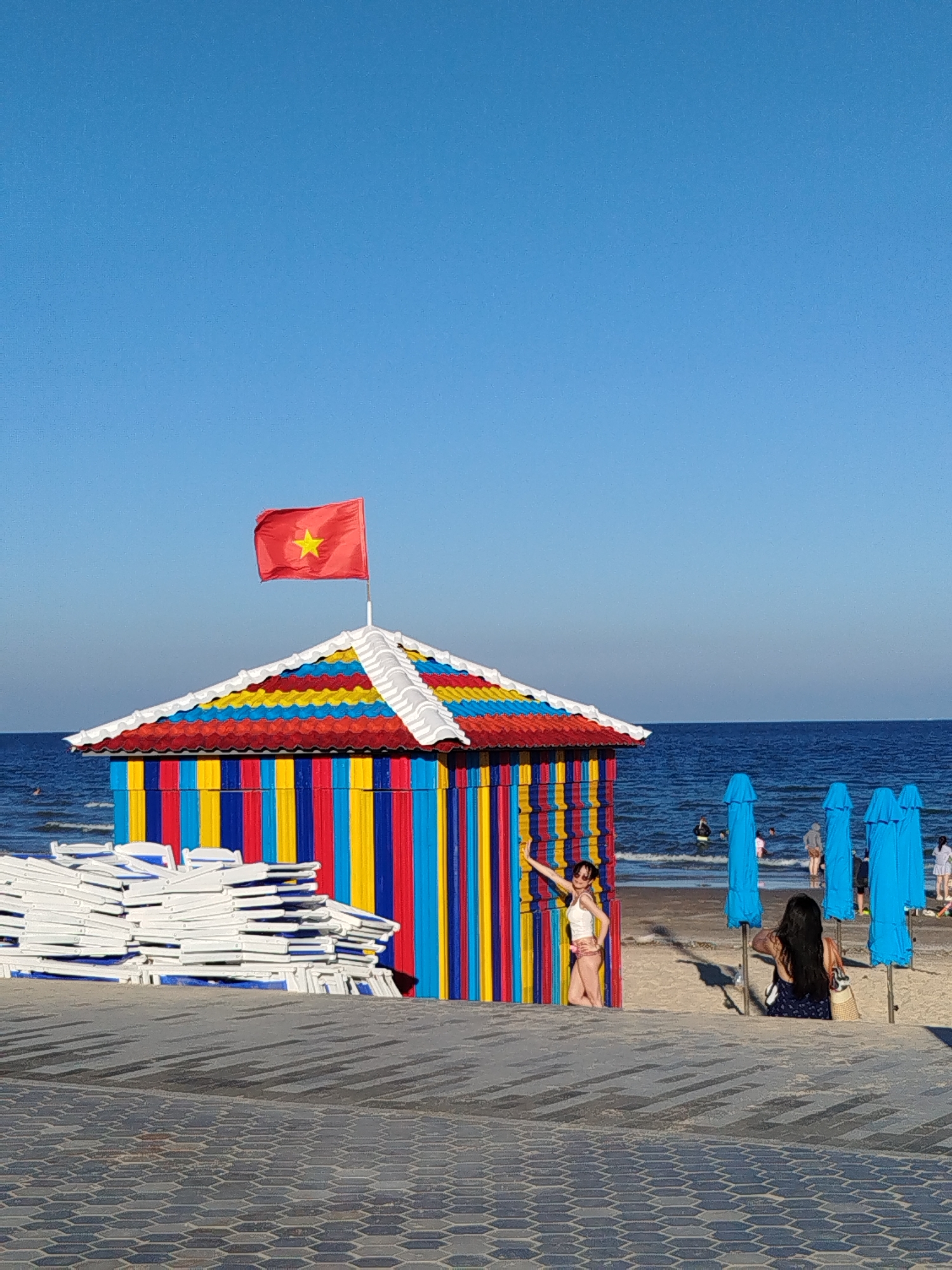 The image captures a beach scene in what appears to be a recreational or tourist area. A small, colorful structure is prominent, featuring vertical stripes of blue, yellow, and red, and a roof with white trim and colored shingles. A Vietnamese flag is mounted on top of the structure. A woman is standing near the structure, seemingly posing for a photo. To the right, another woman is sitting on the sand, facing away from the camera. Blue beach umbrellas line the background. In the foreground, there is a tiled walkway leading to the beach. To the left of the structure, there are stacked white beach chairs. The ocean is visible in the background, with a few people in the water. The sky is clear and blue. Given the presence of the Vietnamese flag and the overall aesthetic, the scene likely takes place in a coastal area near Ho Chi Minh City, Vietnam.