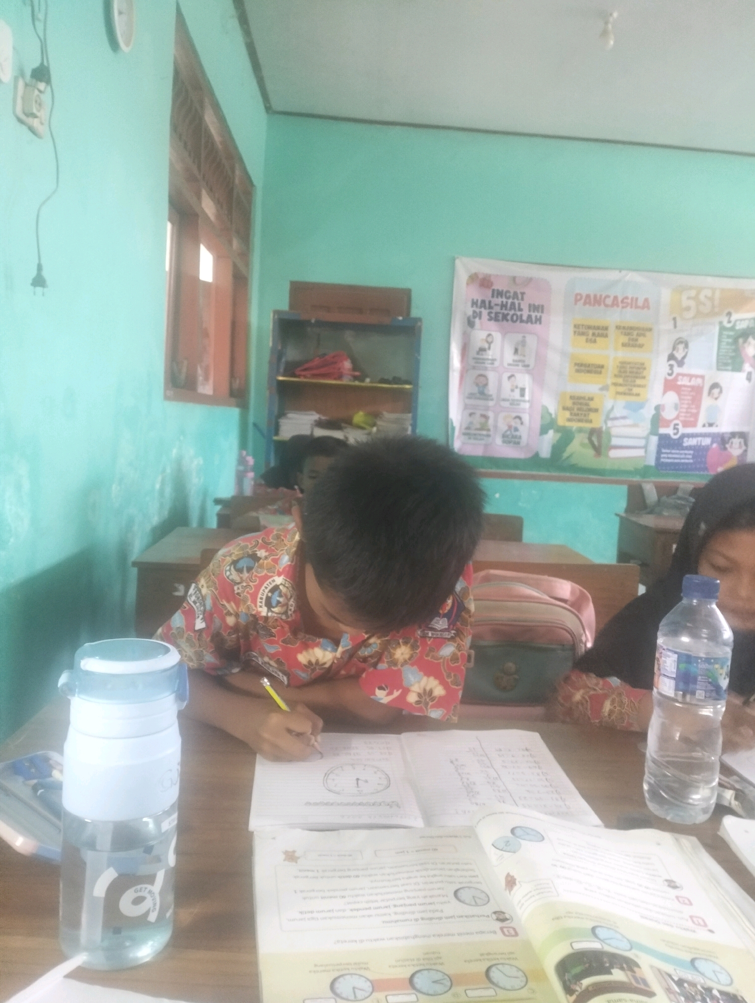 In a classroom in Blabakan, Indonesia, two young students are engaged in learning activities at their desks. The room is painted a light teal color, with a window on the left side featuring wooden blinds. A large poster on the back wall displays educational content with the words "INGAT HAL-HAL INI DI SEKOLAH" (Remember These Things at School) and "PANCASILA" (the state ideology of Indonesia), accompanied by various icons and text in Indonesian.
In the foreground, a boy wearing a red and white batik-patterned shirt is diligently writing in a notebook with a yellow pencil. Beside him, a light blue and white water bottle stands on the table. To his right, a girl with dark hair is also focused on her work, with a bottle of water next to her. Several open books and notebooks are spread across the table, indicating an active learning environment. In the background, shelves are visible, filled with books and other classroom materials. The overall scene depicts a typical day of education in an Indonesian classroom.