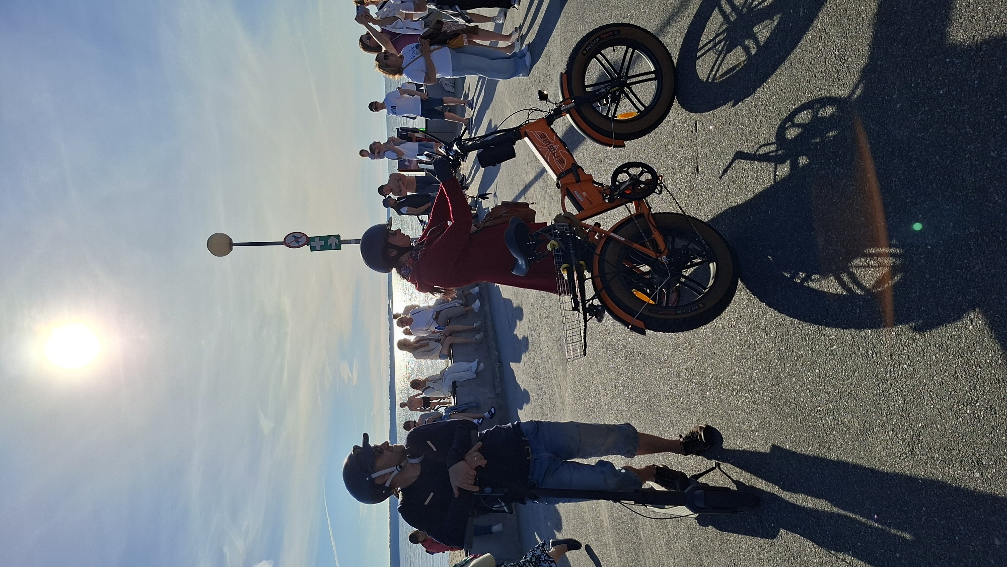 A group of people are gathered by the waterfront in what appears to be a park or promenade. The sun is shining brightly, casting long shadows on the asphalt ground, suggesting it is late afternoon. The sky is clear and blue, with a few wispy clouds.
In the foreground, two individuals are standing. One person, wearing a red garment and a helmet, is near an orange electric bicycle. This bicycle has fat tires and appears to be folded. The second person, also wearing a helmet, is in a black top and denim shorts, standing next to another bicycle which is mostly out of frame.
Further back, a line of people are sitting on a low wall overlooking the water, their backs to the camera. They are dressed in casual summer clothing. Behind them, more people are standing and walking, some holding phones as if taking pictures. The scene suggests a relaxed, social gathering, possibly a tourist spot or a popular leisure area.
A lamppost with two signs is visible. One sign shows a red circle with a diagonal line through it, indicating a prohibition, and a green arrow pointing upwards. The other sign appears to be a green exit sign with an arrow. The presence of the water and the clear sky indicate an outdoor setting, likely a coastal area. The overall mood seems to be pleasant and leisurely, with people enjoying the sunny weather. The text "RIDEN STAR" is visible on the tires of the orange bicycle.
