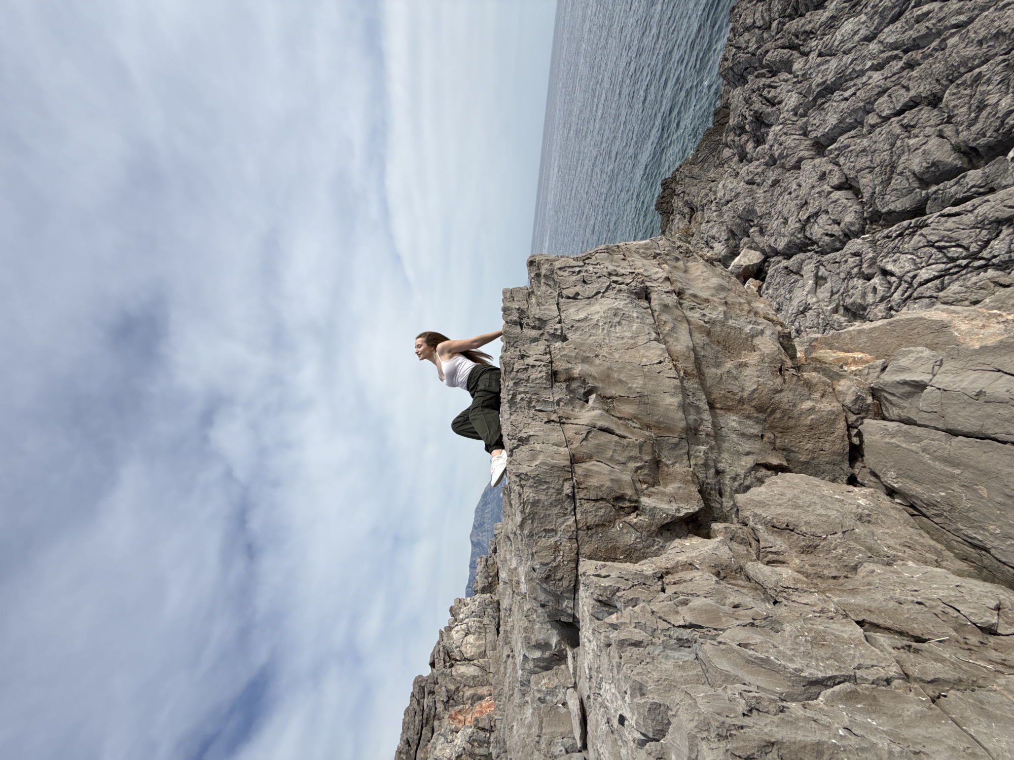 A young woman in a white tank top, dark pants, and white shoes is leaning off the edge of a rocky cliff. She is gripping the rock face with both hands. The backdrop shows a cloudy sky, transitioning to the sea at the upper-right. The location is Boreti, Montenegro.