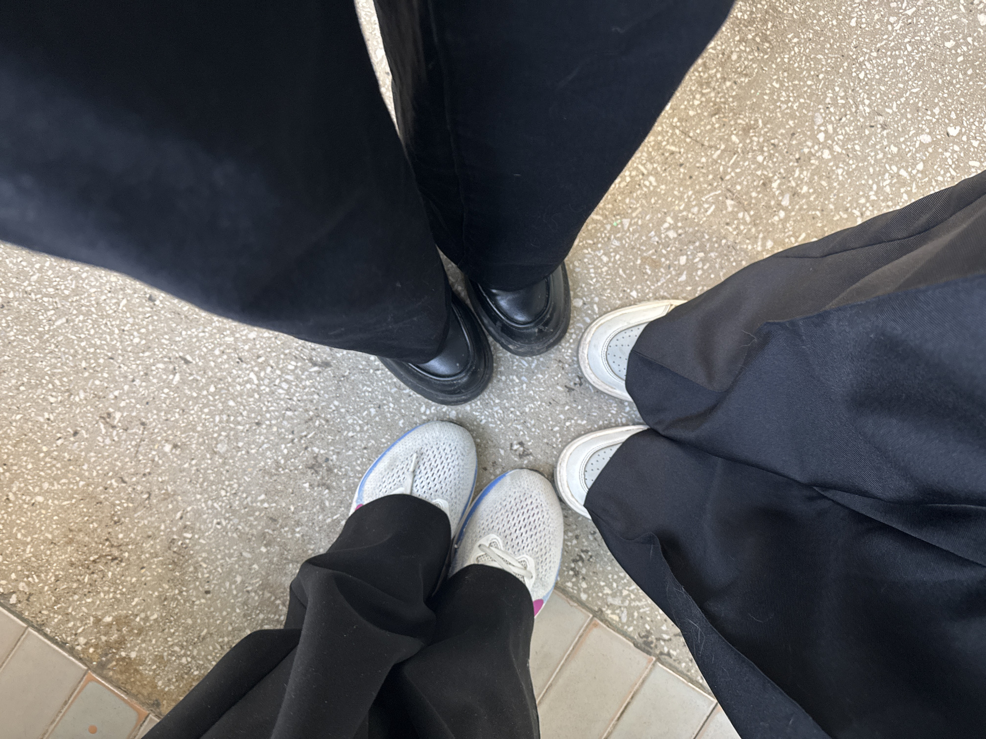 The image presents an overhead view of the lower legs and feet of three individuals, forming a circular arrangement on a speckled floor. All three subjects are wearing dark, likely black, trousers.
One pair of feet, positioned at the top of the image, wears black, possibly leather, loafers with thick soles. The trousers appear to be made of a textured, uniform-like material.
Another pair of feet, on the bottom left, sports light grey mesh sneakers with white laces and subtle blue and pink accents visible on the sides. These are paired with flowing black trousers.
The third visible pair of feet, on the right, wears white sneakers with grey perforated sections and white laces, also with dark trousers that appear to be made of a smooth, slightly textured fabric.
The ground beneath them is a light gray and beige speckled surface, possibly concrete or terrazzo, with a border of larger, lighter square tiles visible in the bottom-left corner. The lighting is bright and even, suggesting daytime, likely indoors or in a well-lit covered outdoor area. The gathering appears casual, with the individuals standing close together, perhaps conversing. There is no visible text in the image. The setting is in Chaikovsky, Russia.