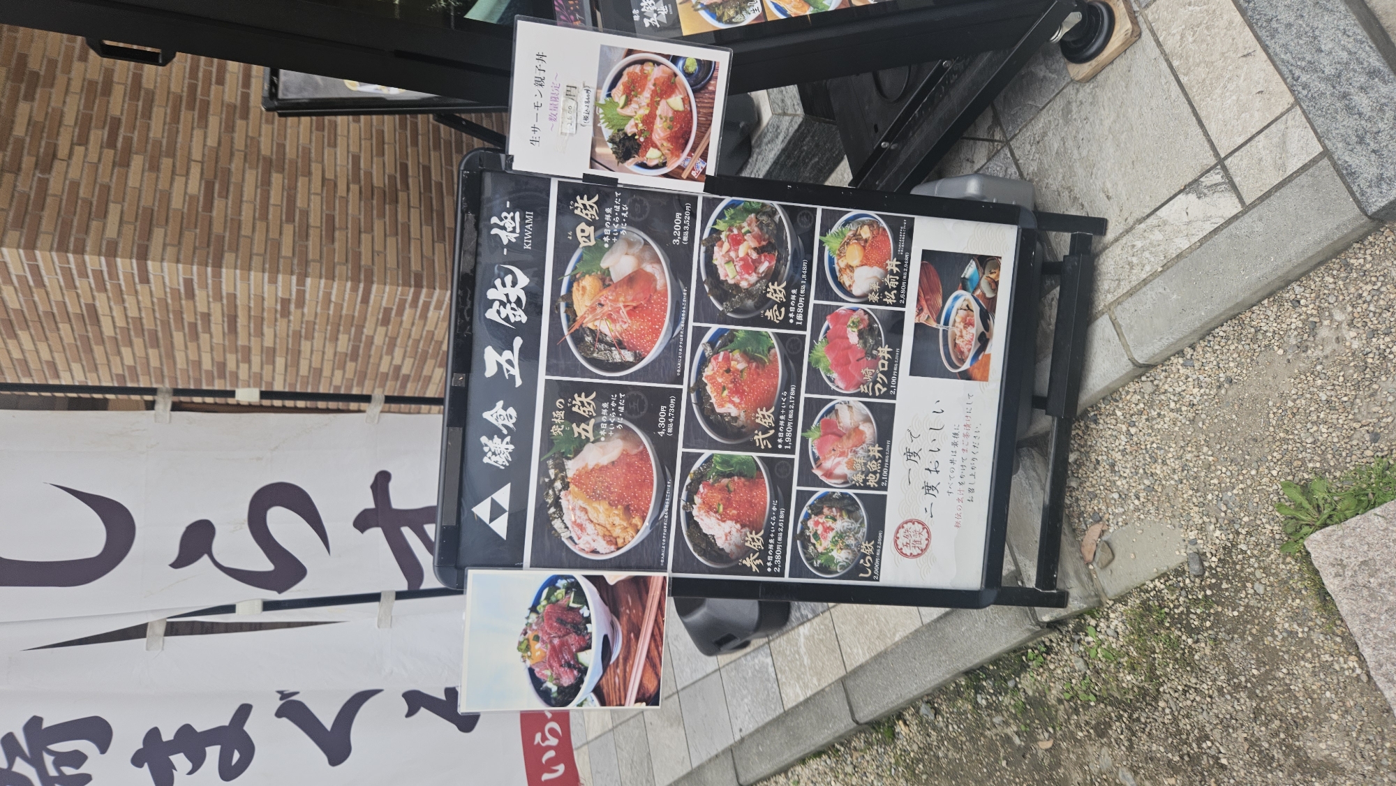 The image shows an outdoor menu board for a restaurant called "鎌倉 五鉄 -極-" (Kamakura Gotetsu -Kiwami-). The board displays various seafood rice bowls with prices. There are multiple images of the dishes, each with a Japanese name and price. The menu items include "究極の五鉄" (Ultimate Gotetsu), "海鮮四鉄" (Seafood Tetsu), "参鉄" (San Tetsu), "武鉄" (Take Tetsu), "壱鉄" (Ichi Tetsu), "海鮮地魚丼" (Seafood Local Fish Bowl), "三崎マグロ丼" (Misaki Tuna Bowl), and "松前丼" (Matsumae Bowl). A separate small sign on the right side advertises "生サーモン親子丼" (Raw Salmon and Roe Bowl) for 2,600 yen. The menu board is placed on a sidewalk next to a brick wall. The ground is paved with concrete and some gravel. The overall scene suggests an advertisement for a restaurant in Kamakura, Japan, likely in a public area where people can view the menu before entering.