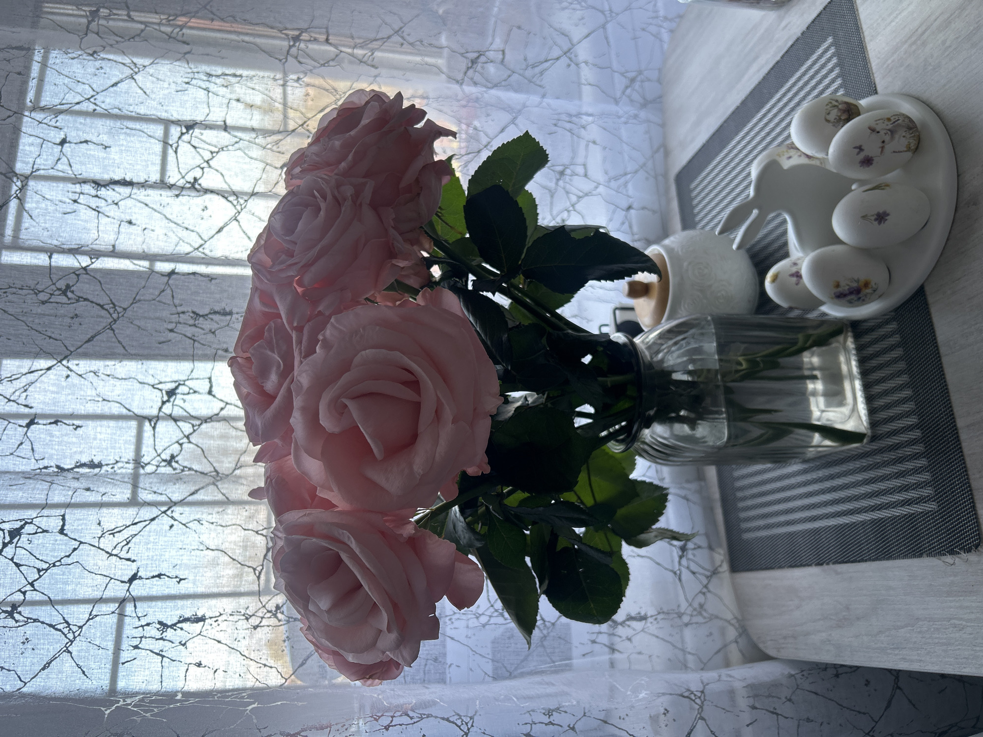 The image shows a bouquet of pink roses in a glass vase. The roses are the main focus and are arranged with green leaves. Behind the roses, there's a patterned white curtain, slightly obscuring a window with a grid-like structure. To the right of the vase, there is a decorative arrangement that includes an egg holder, small painted eggs, and possibly a small ceramic rabbit on a placemat. The surface on which the arrangement rests appears to be a table with a marble-like pattern. The scene is presumably indoors, lit by natural light coming through the window. There's no visible information to confirm the location is in Klin, Russia.