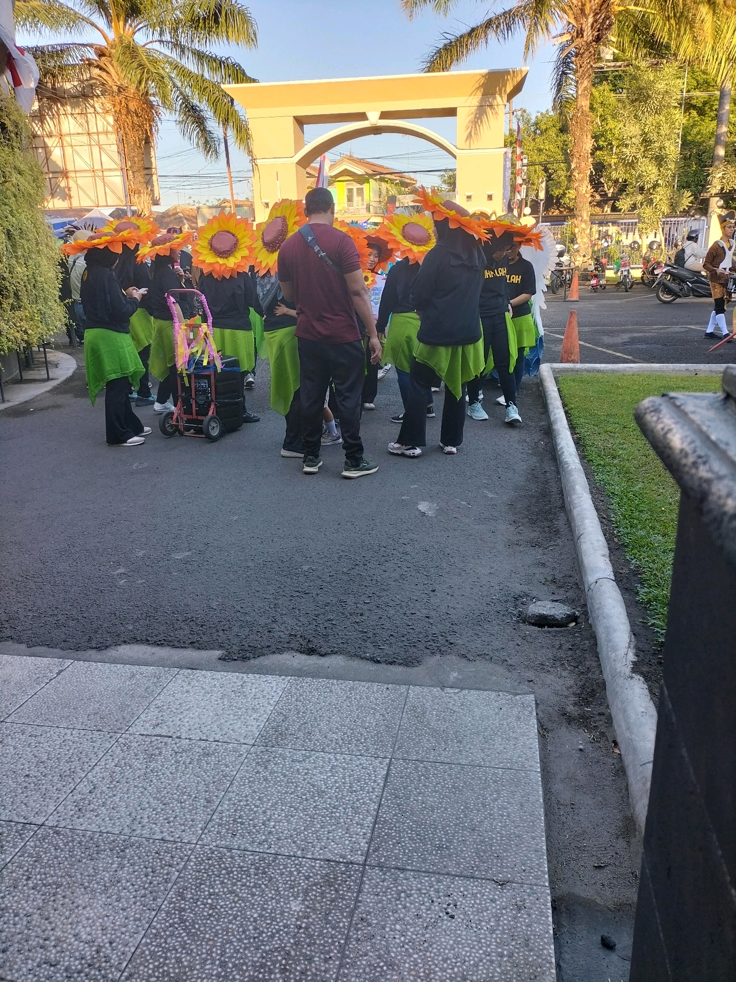 In Pare, Indonesia, a group of people, primarily women, are gathered in a street lined with palm trees and a large arched entrance structure. They are dressed in matching black tops and bright green skirts, with many wearing oversized sunflower-shaped hats. One individual, a man wearing a maroon t-shirt and black pants, stands out among the group. A small wheeled cart, decorated with colorful ribbons and streamers, is positioned in the center of the gathering. The scene appears to be part of a festive event or parade, with a clear blue sky suggesting daytime. The ground is paved with asphalt and bordered by a concrete curb and a grassy area. In the background, buildings and parked motorcycles are visible, indicating an urban or semi-urban setting.