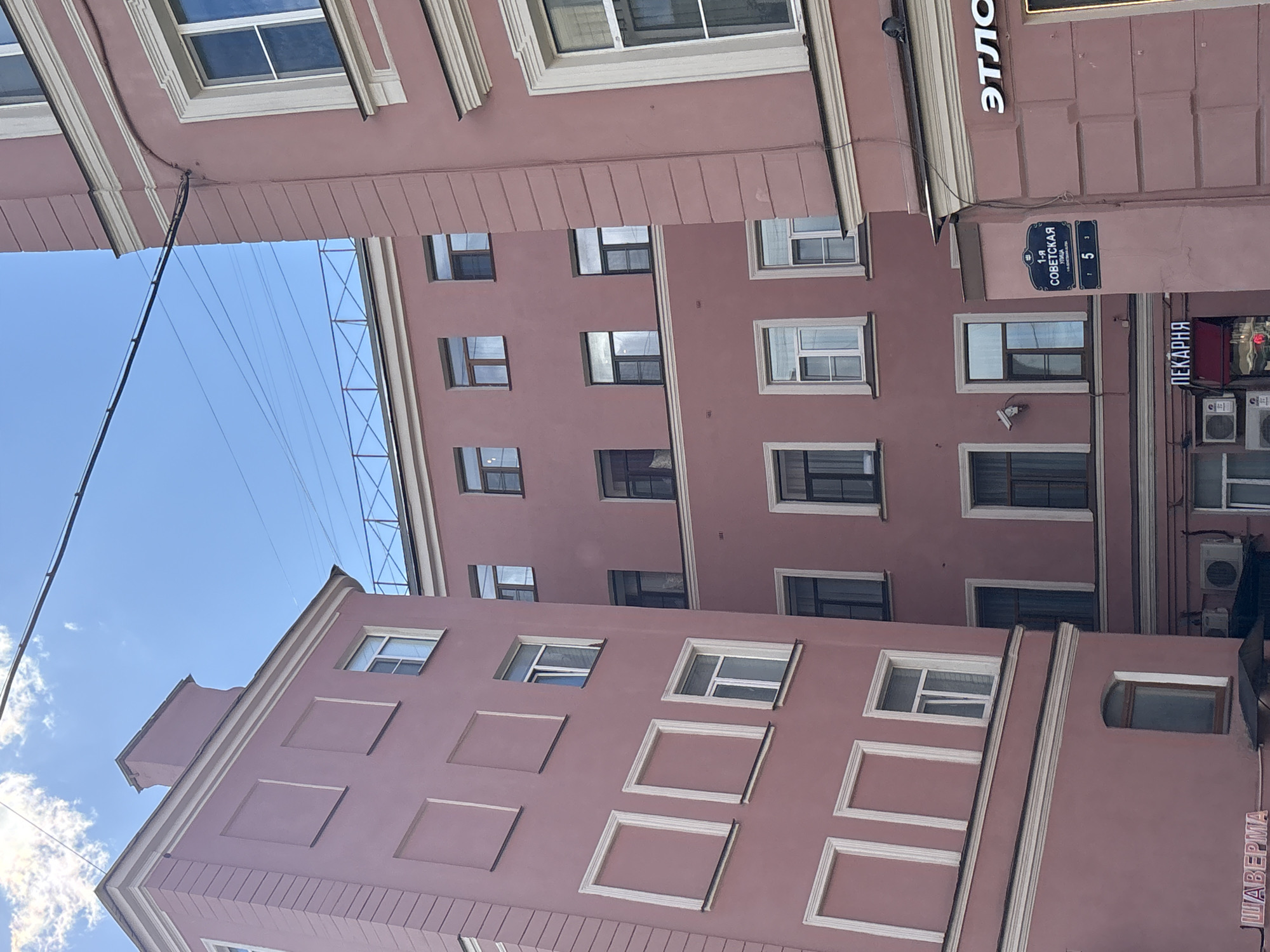 The image shows a view of buildings with pink facades in Saint Petersburg, Russia, under a bright blue sky with some clouds. The architecture features multiple windows, some with decorative white frames, and layered cornices. A street sign is visible on a cylindrical column, indicating "1-я СОВЕТСКАЯ УЛИЦА" (1st Soviet Street), with the number "5". Other signs in Russian are also visible, including "ПЕКАРНЯ" (Bakery) and "ЭТЛО" (possibly part of a store name like "Этюд"). Electrical wires are strung across the sky. The buildings appear to be residential or mixed-use. The lighting suggests it is daytime.