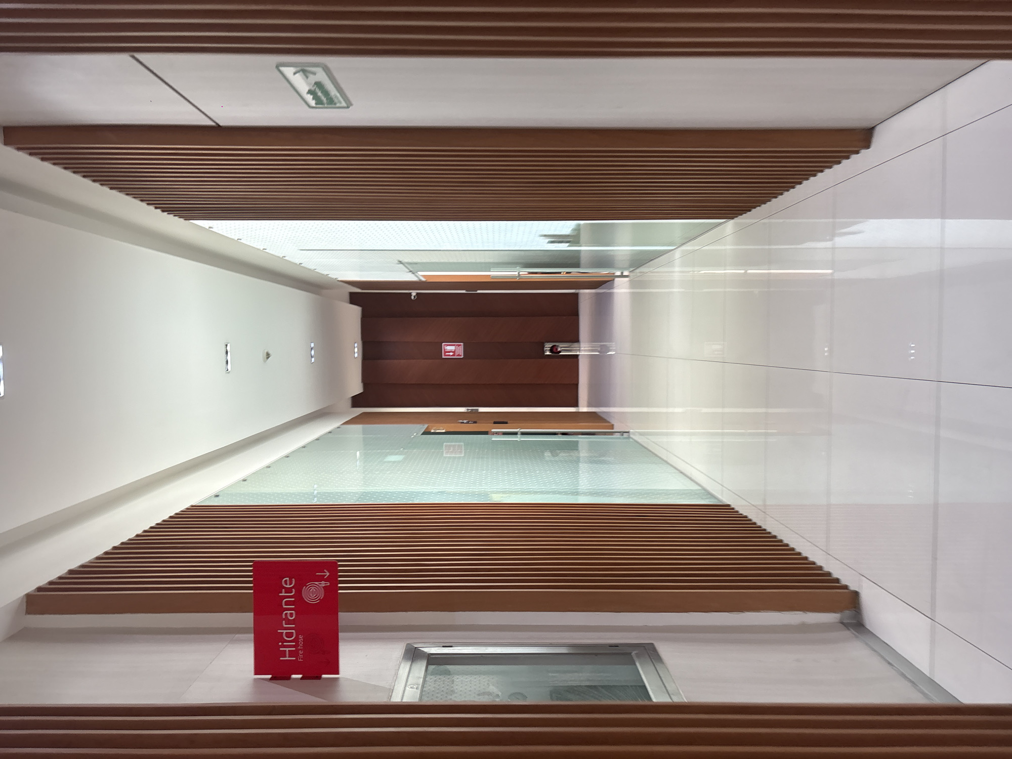 A view of a modern building's interior, likely a commercial or institutional space. The scene is organized as a corridor. On the left, the walls are white with flush, rectangular indentations. The right wall is covered with large, square, white tiles. Wooden slats form a parallel pattern along the top and bottom of the scene, potentially serving as ceiling or floor features. Glass walls or partitions are visible halfway down the corridor, creating a view to the floor below. The building features a red sign with the word "Hidrante" and an arrow. At the end of the corridor is a brown wall with a red fire alarm, and a figure is faintly visible in the distance. The photograph appears to be taken in the city of San Bernardino Tlaxcalancingo, Mexico, based on the presence of Spanish-language signage.